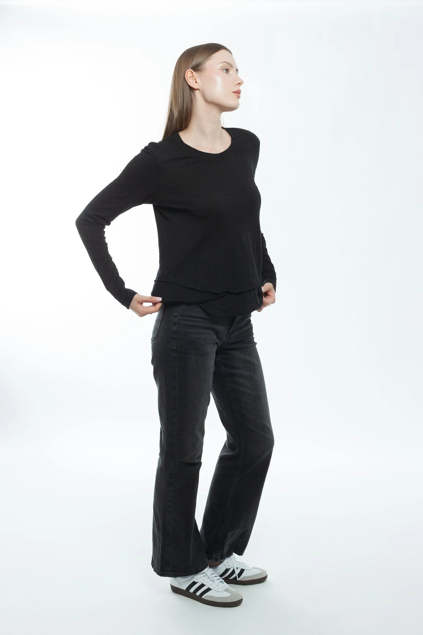 A young woman with long hair wearing a black long-sleeve top, dark jeans, and white sneakers, standing against a plain white background.