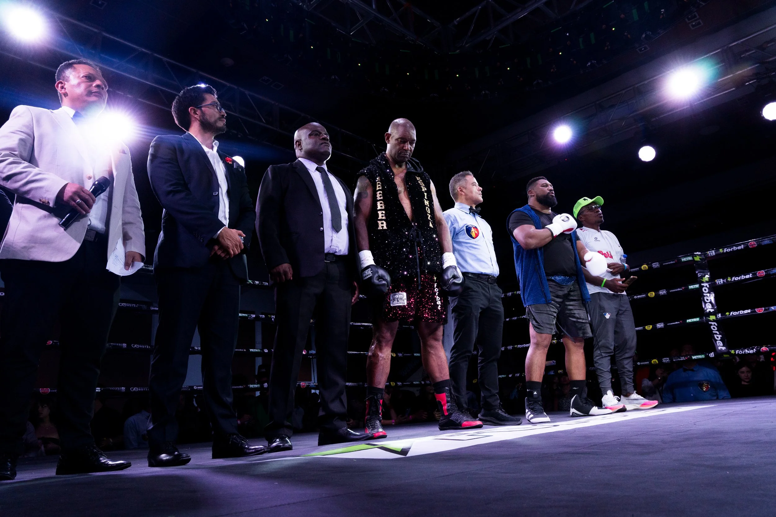 Boxers and officials standing in a ring before a match, with bright lights overhead and an audience in the background.