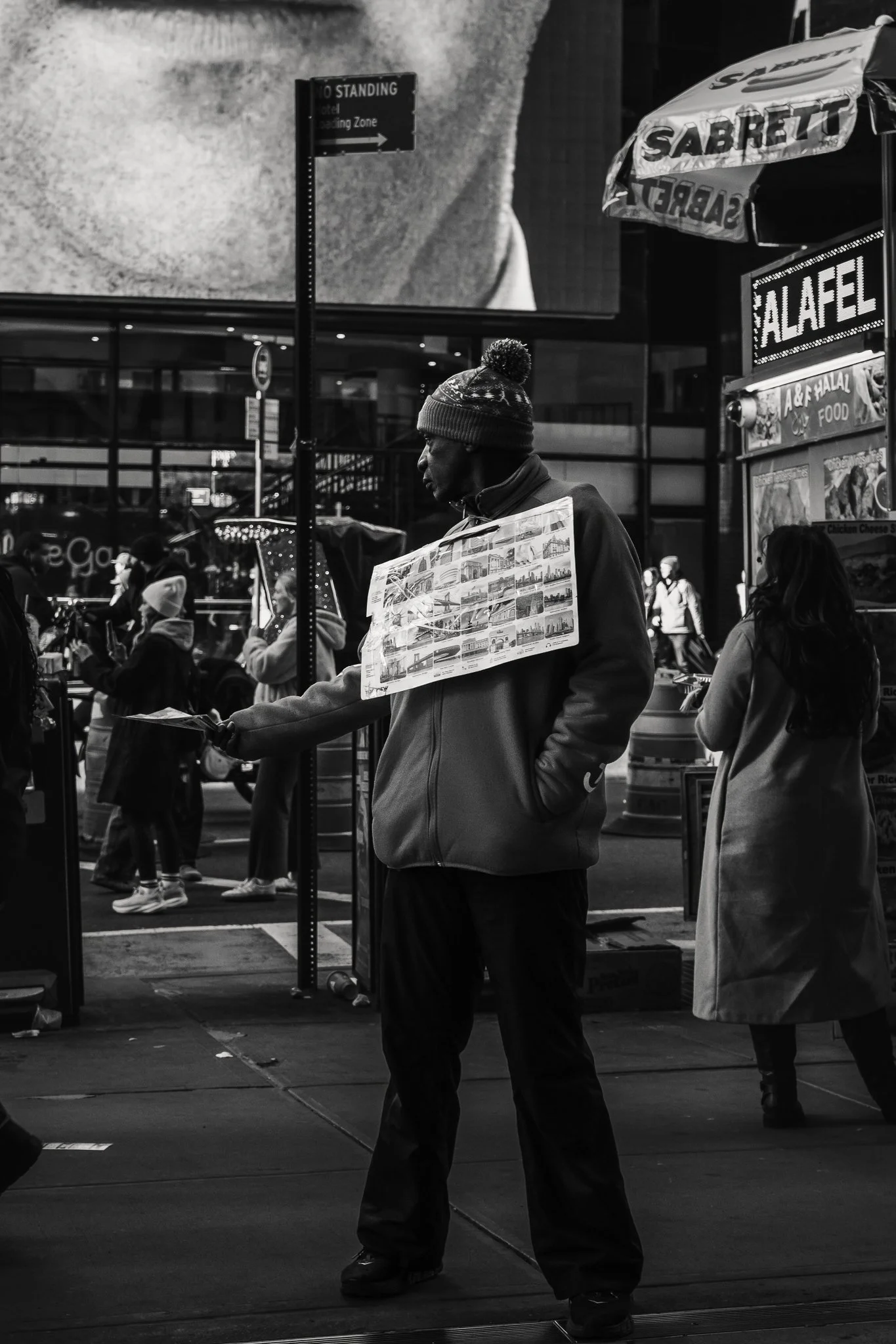 A man wearing a knit cap and hoodie standing on a city sidewalk at night, holding some papers or flyers. There are other pedestrians in the background, and signs and storefronts are visible, indicating a busy urban area.