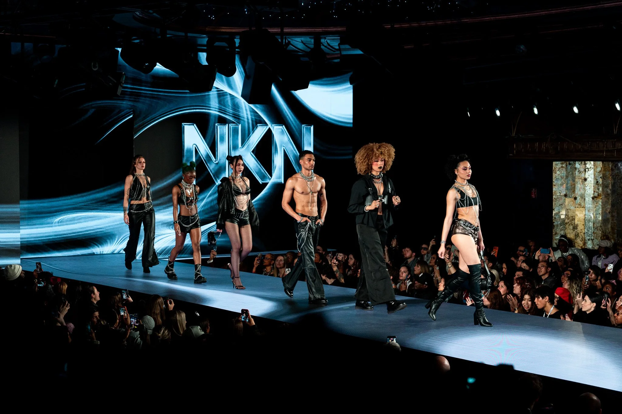 Models walking down a runway during a fashion show, with an audience watching and taking photos. The background features the