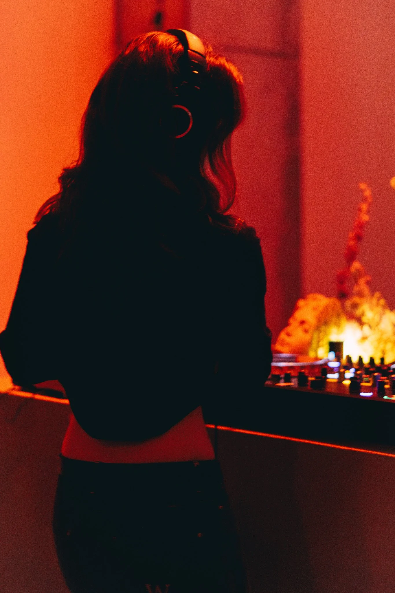 A woman with long hair wearing headphones standing in a dimly lit room with red lighting, next to DJ equipment with colorful lights and flowers in the background.
