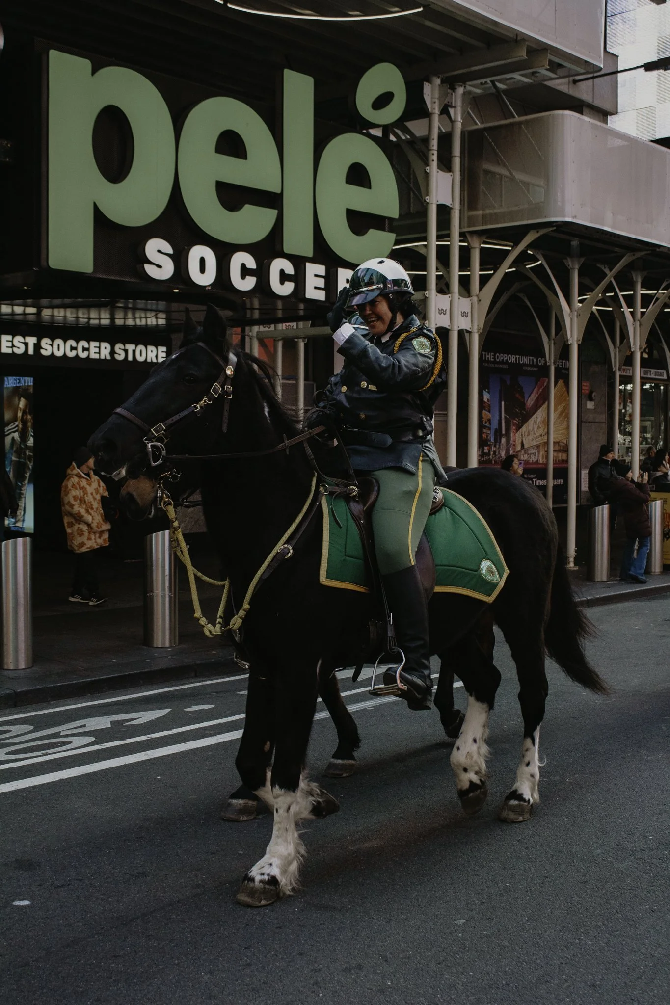 A police officer riding a horse on a city street in front of a large sign for 'Pelle Soccer'. The horse is black with white markings on its legs and face, and the police officer is smiling and talking, wearing a helmet and uniform. People are walking
