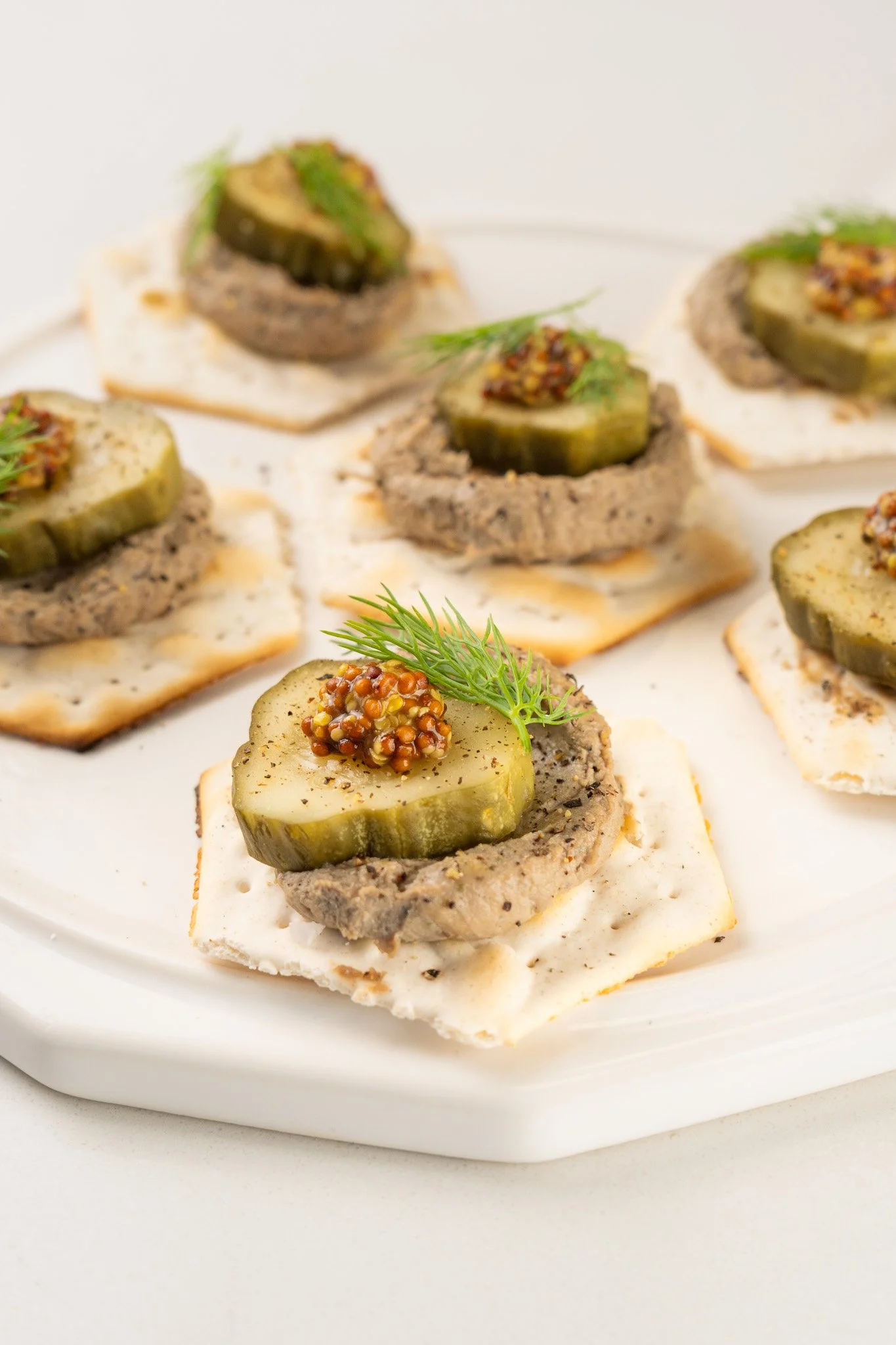 Appetizer pieces with pickles, mustard, and herbs on crackers on a white plate.