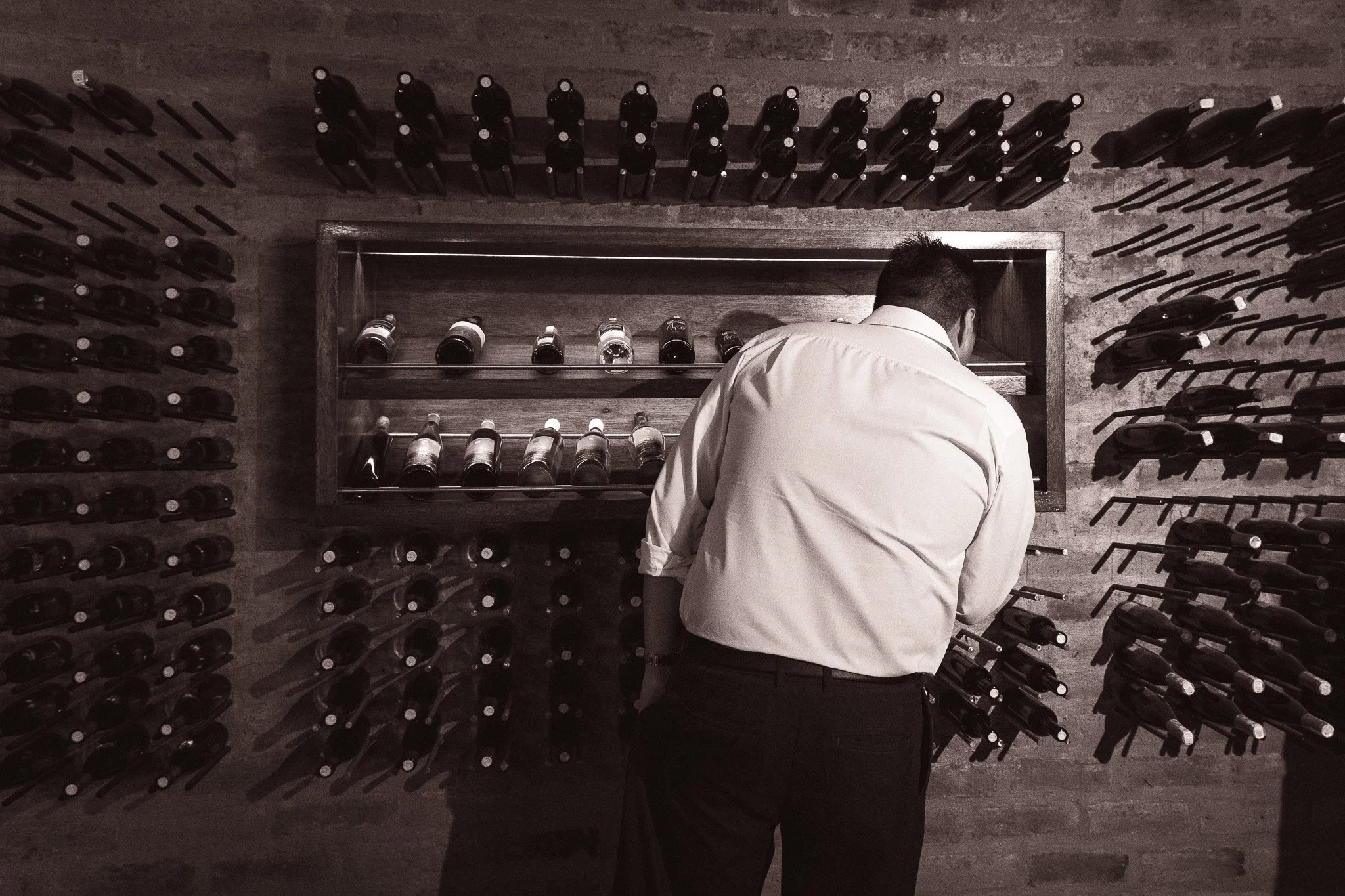 A man in a white shirt is looking at a wine bottle collection on dark wood shelves in a wine cellar or bar, with multiple rows of wine bottles on the walls around him.