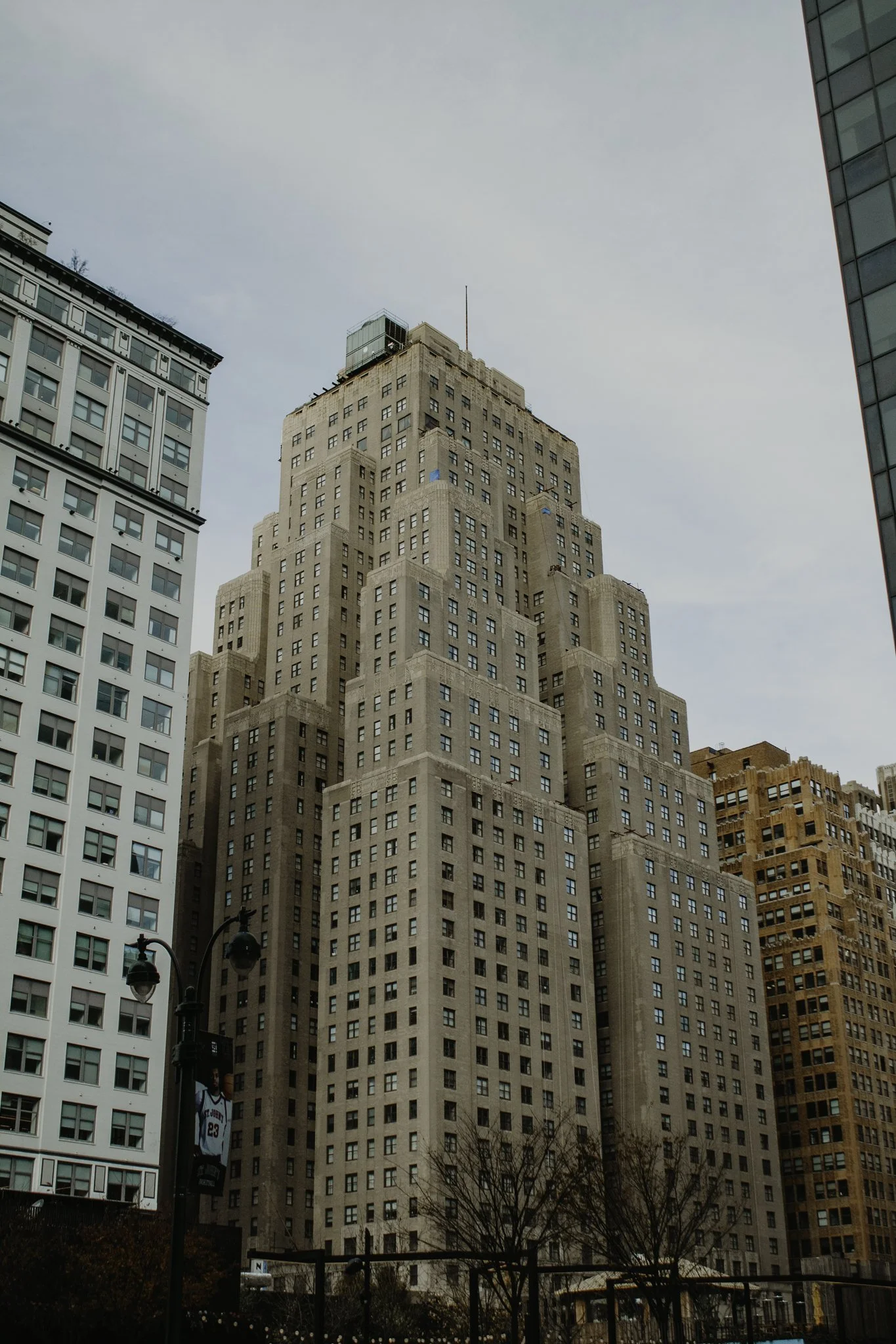 Tall granite skyscraper with Art Deco architecture, surrounded by modern office buildings, streetlights, and leafless trees in a city setting during daytime.