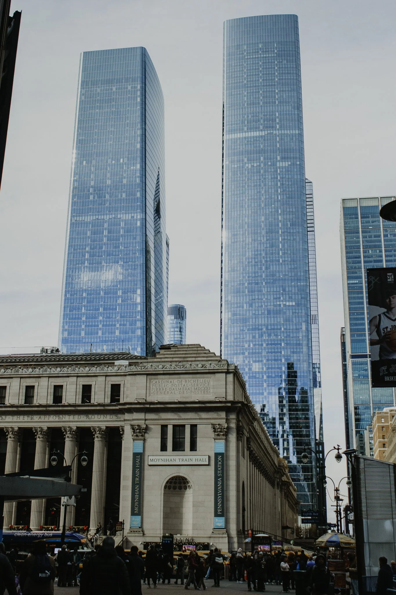 View of New York City Penn Station with modern high-rise buildings in the background, featuring reflective glass facades, and busy pedestrians and yellow taxis in the foreground.