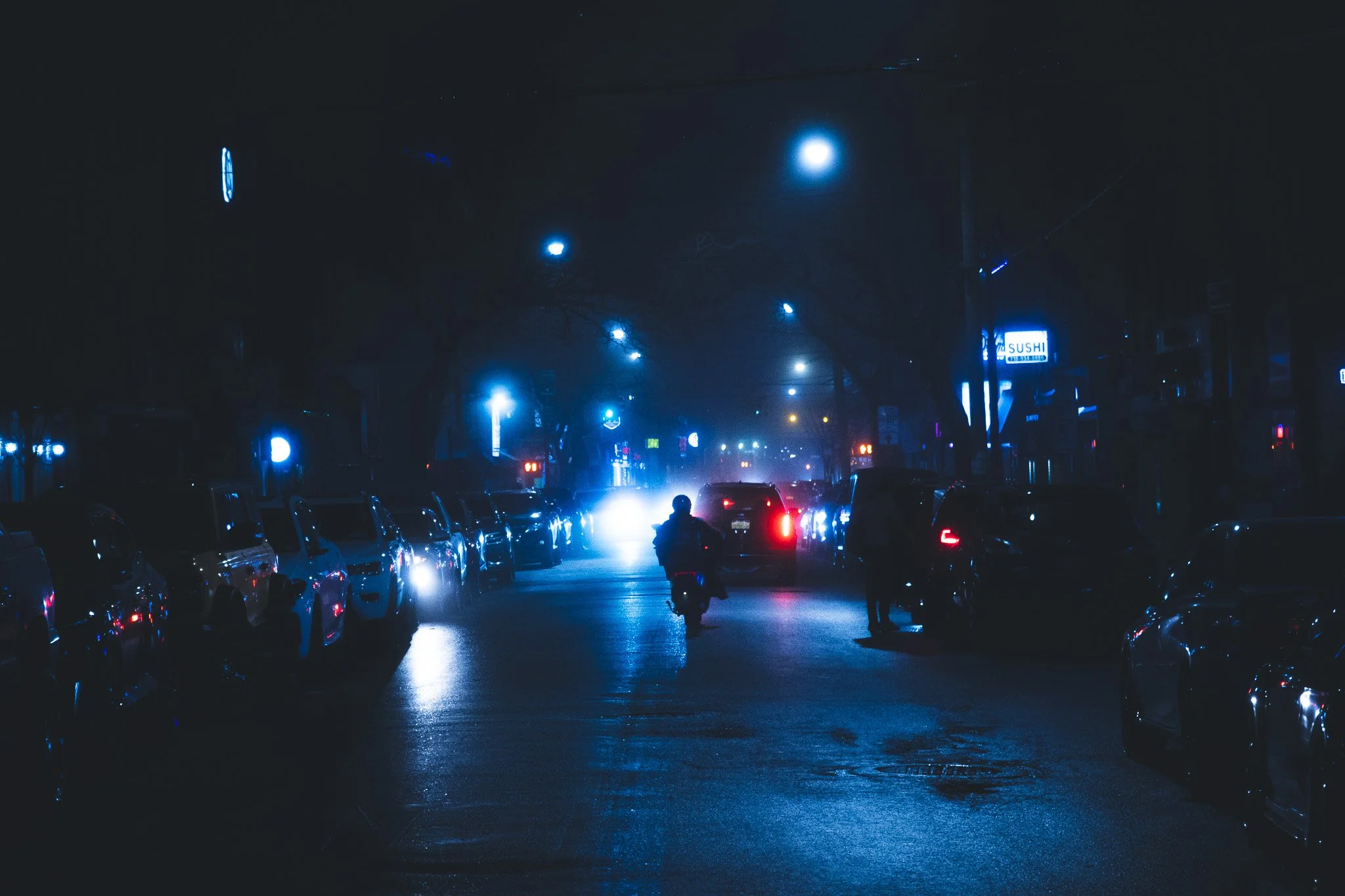 Night street scene illuminated by blue streetlights, with parked cars along the sides, a motorcycle, and a person walking or riding in the street. Buildings with neon signs and a sushi restaurant sign are visible in the background.