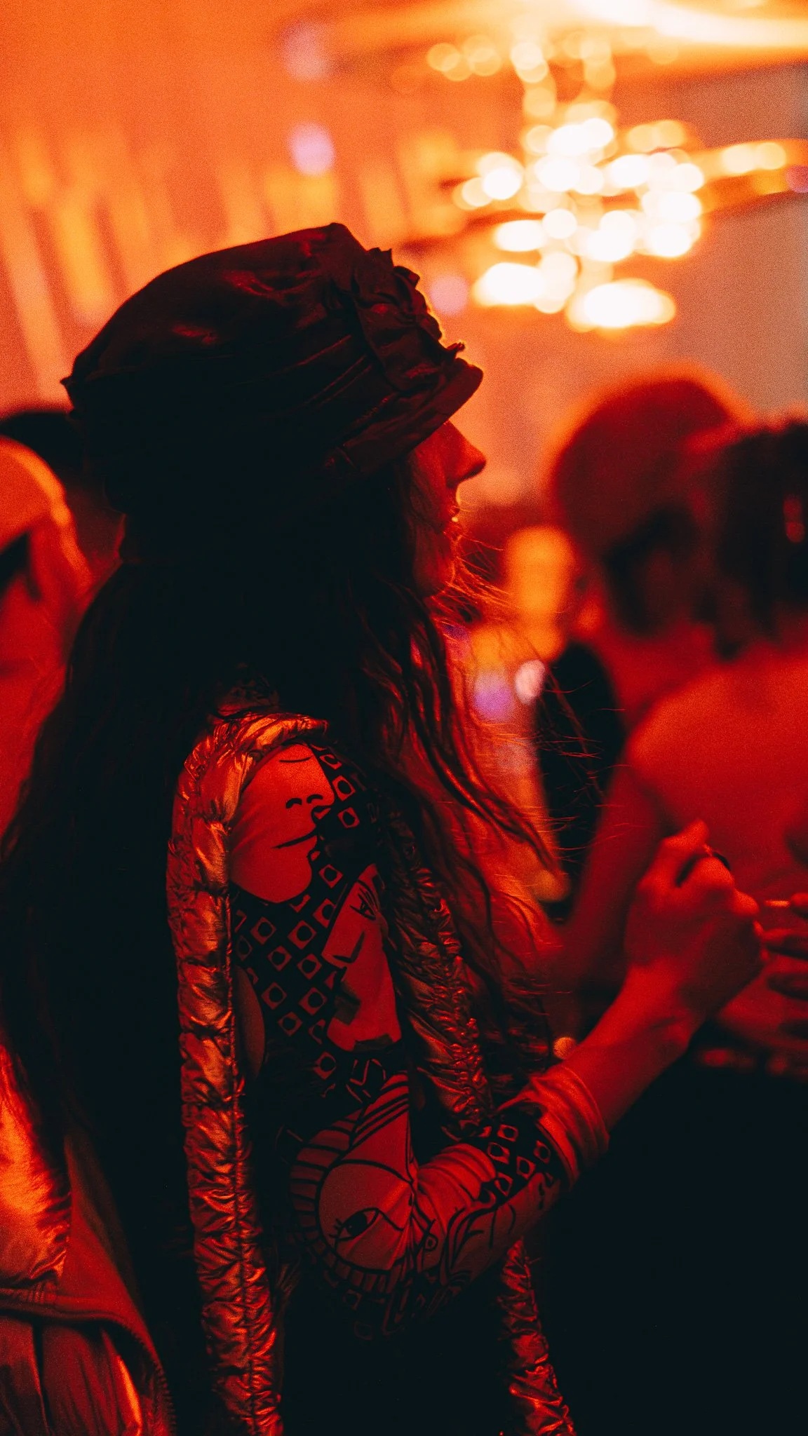 A woman with long hair, wearing a headscarf, is at a gathering or event with colorful, warm lighting, surrounded by other people.