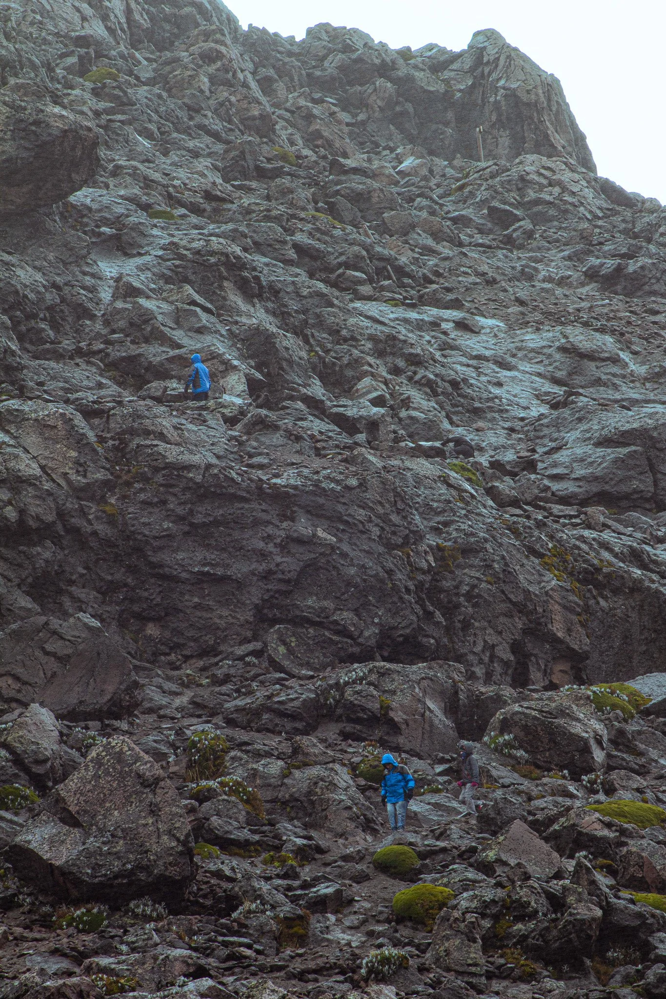 People hiking on a rocky, mountainous terrain with steep, jagged rocks and boulders, some covered with moss, under a cloudy sky.
