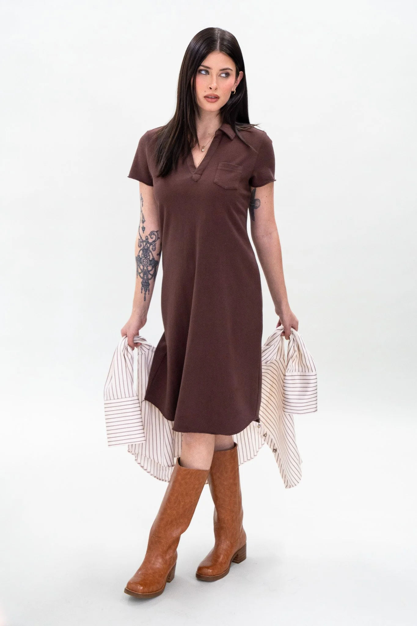A woman with long dark hair, tattoos on her left arm, wearing a brown dress, holding an off-white striped shirt, standing in front of a plain white background.
