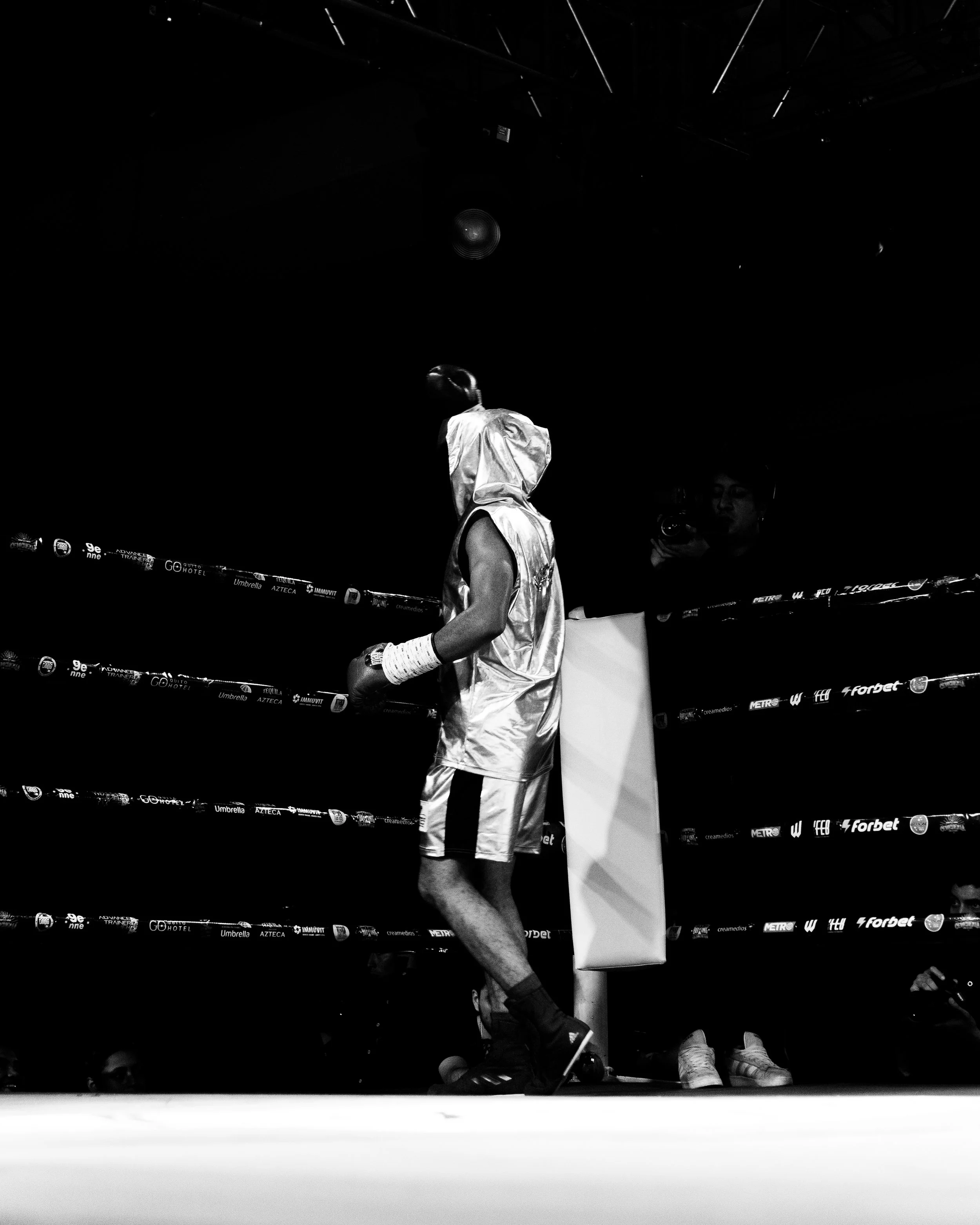 A boxer in white shorts and gloves stands in a boxing ring, with a person crouching on the floor and another person in the background, illuminated by overhead lights in an indoor setting.