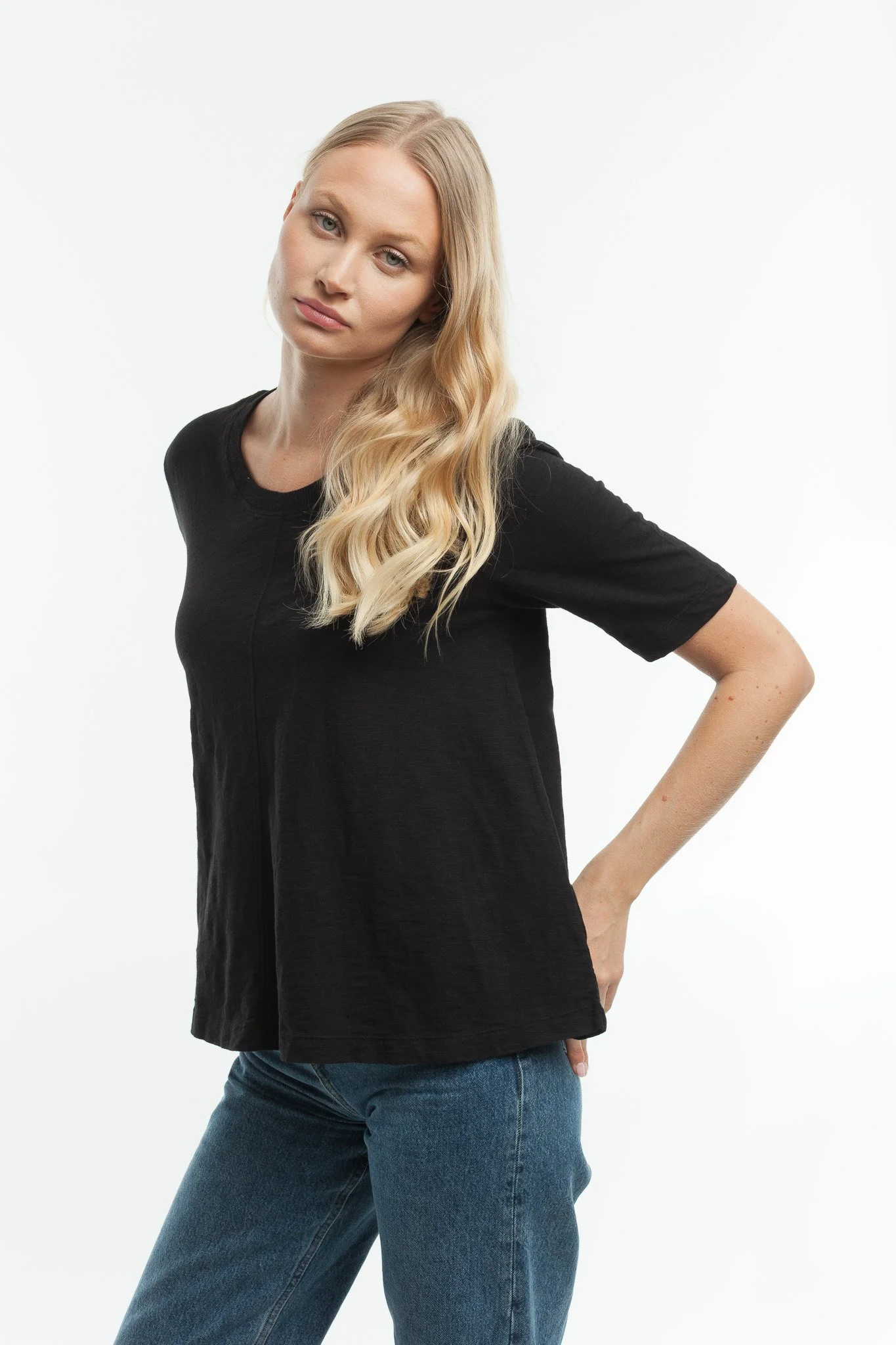 A young woman with long blonde hair in a black t-shirt and blue jeans stands against a white background.