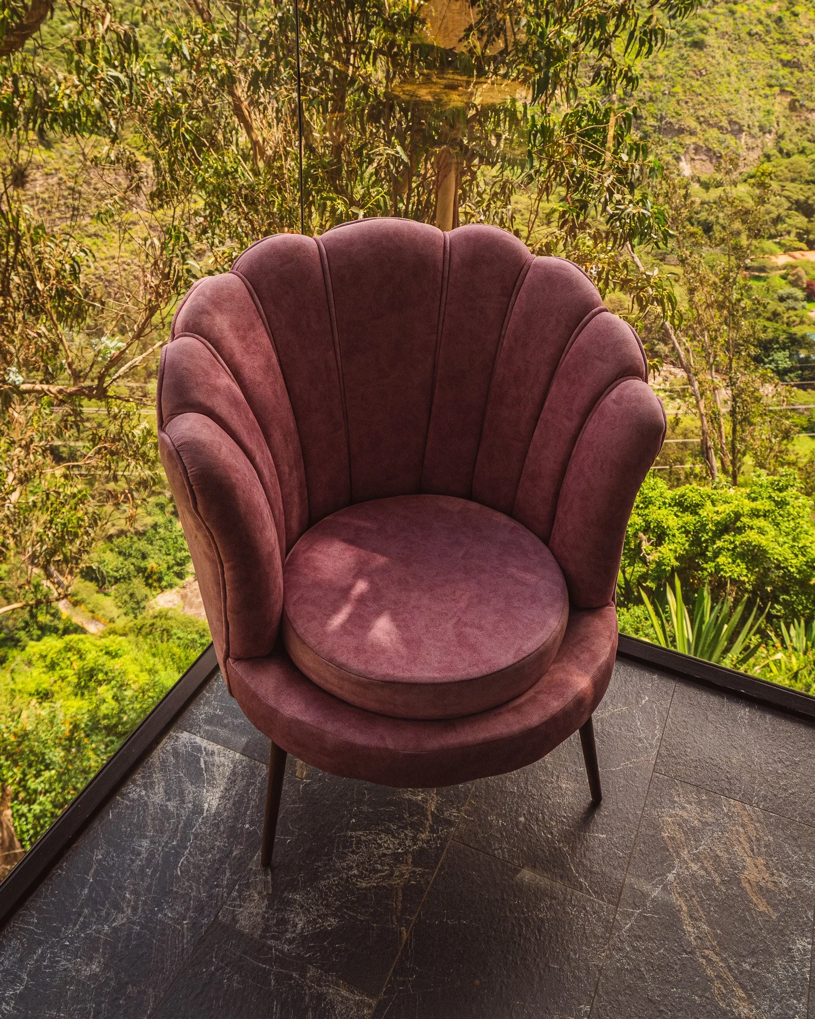 An elegant pink velvet chair with a rounded backrest in front of a large window showcasing a lush green mountain landscape.