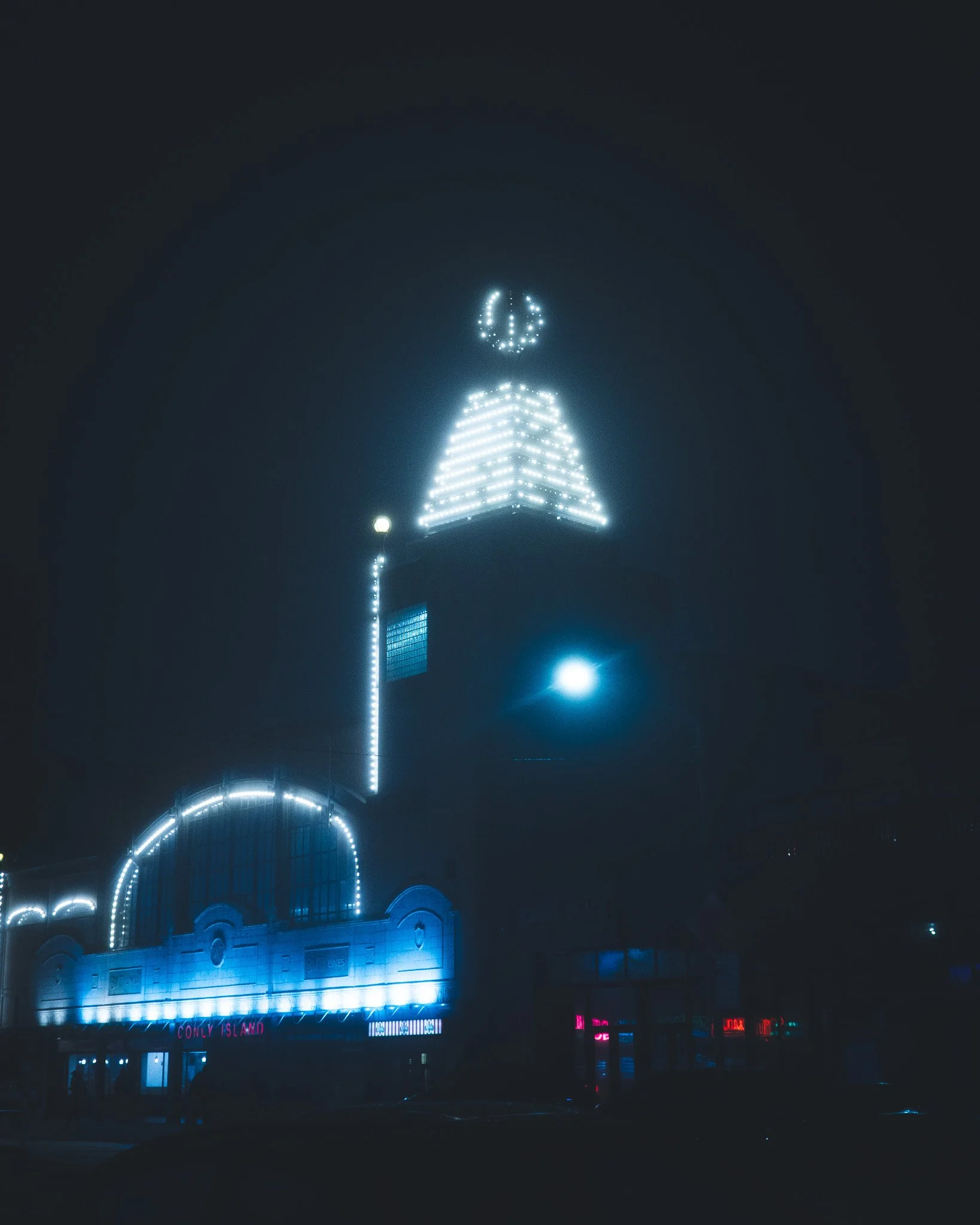 Night view of a building with bright neon lights outlining its architectural features, including a pointed roof and a large clock at the top, with colorful illuminated signs at the bottom.