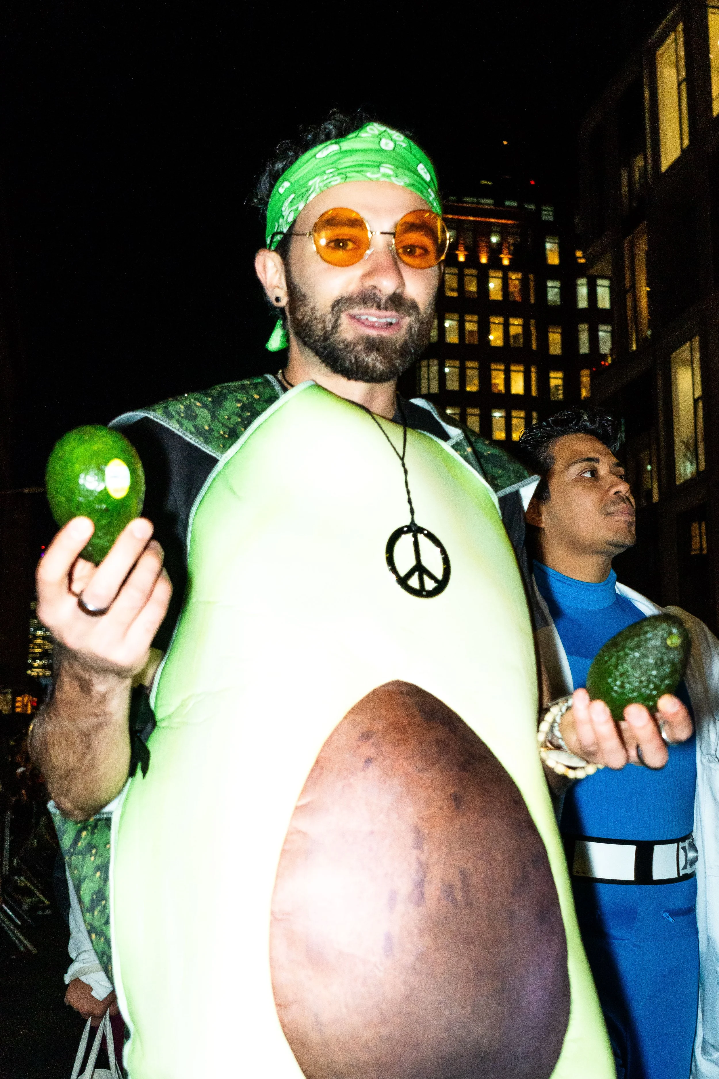 Person dressed as an avocado, wearing sunglasses and a green bandana, holding two avocados, standing at night in an urban area with illuminated buildings in the background.