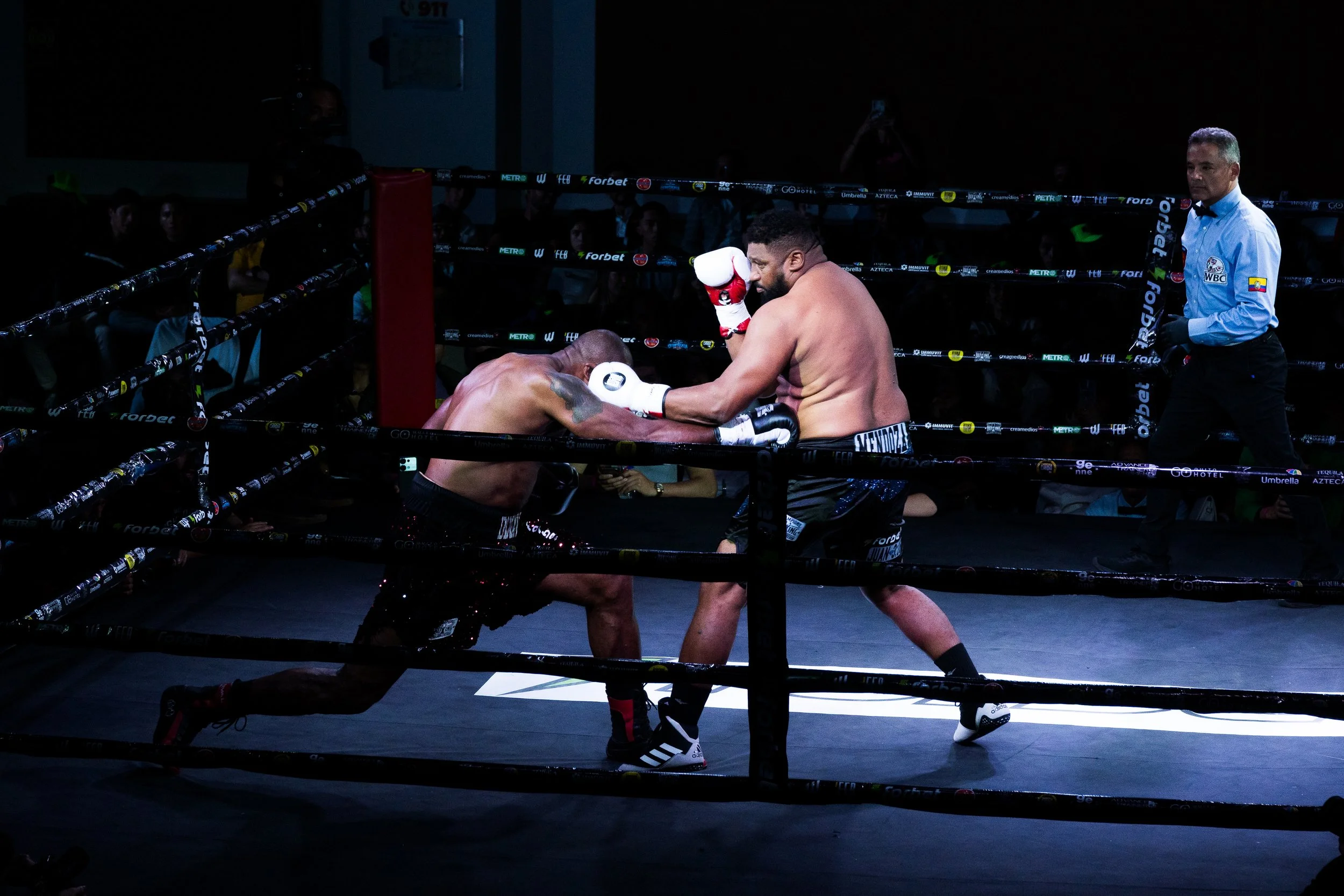 Two boxers fighting in a dark boxing ring, with a referee observing them.