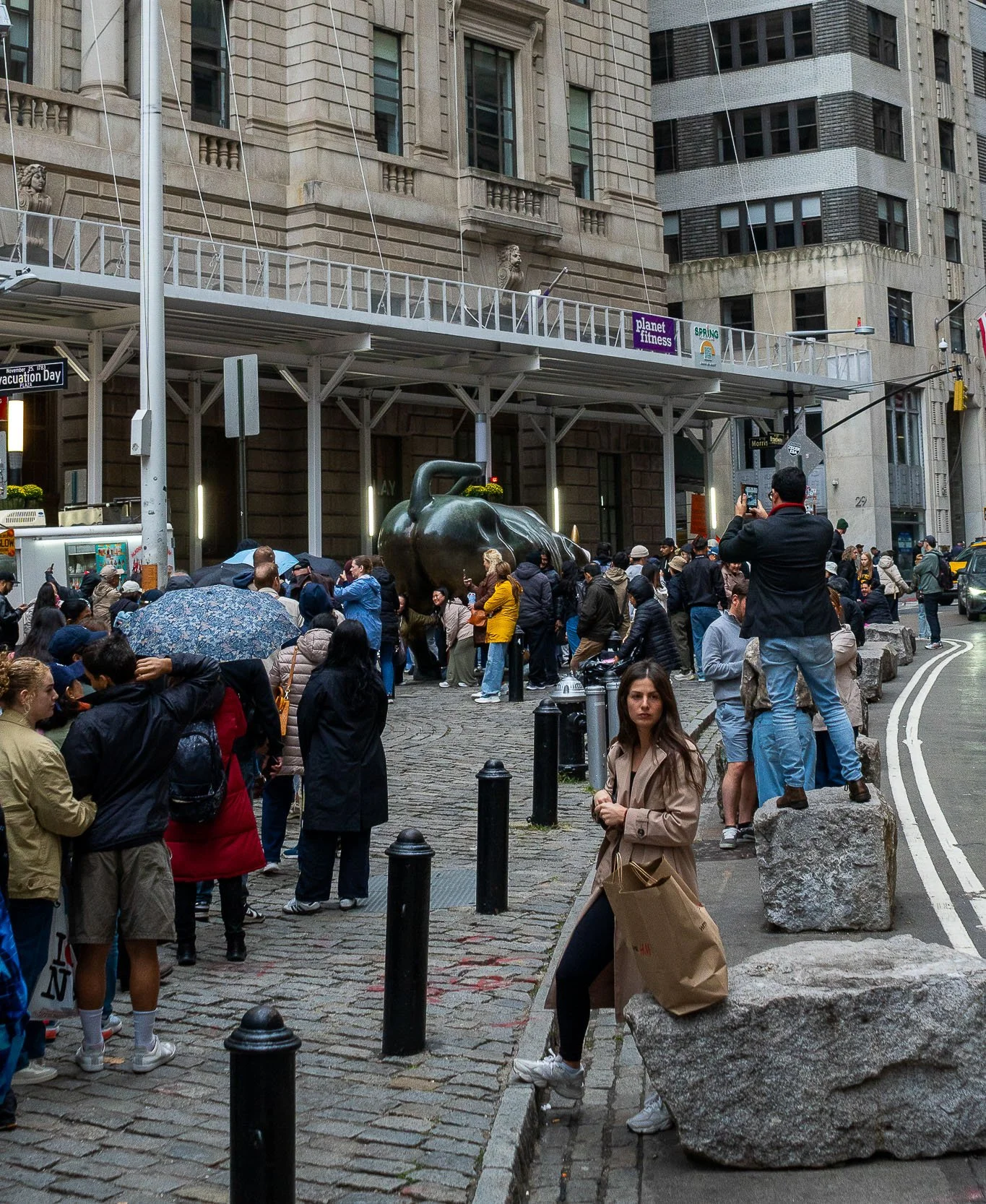 Crowded city street scene with people waiting in line, some taking photos. A large bronze sculpture of a pig is in the center. Surrounding buildings have signage, including Planet Fitness and Spring. The street has cobblestones and large rocks servin