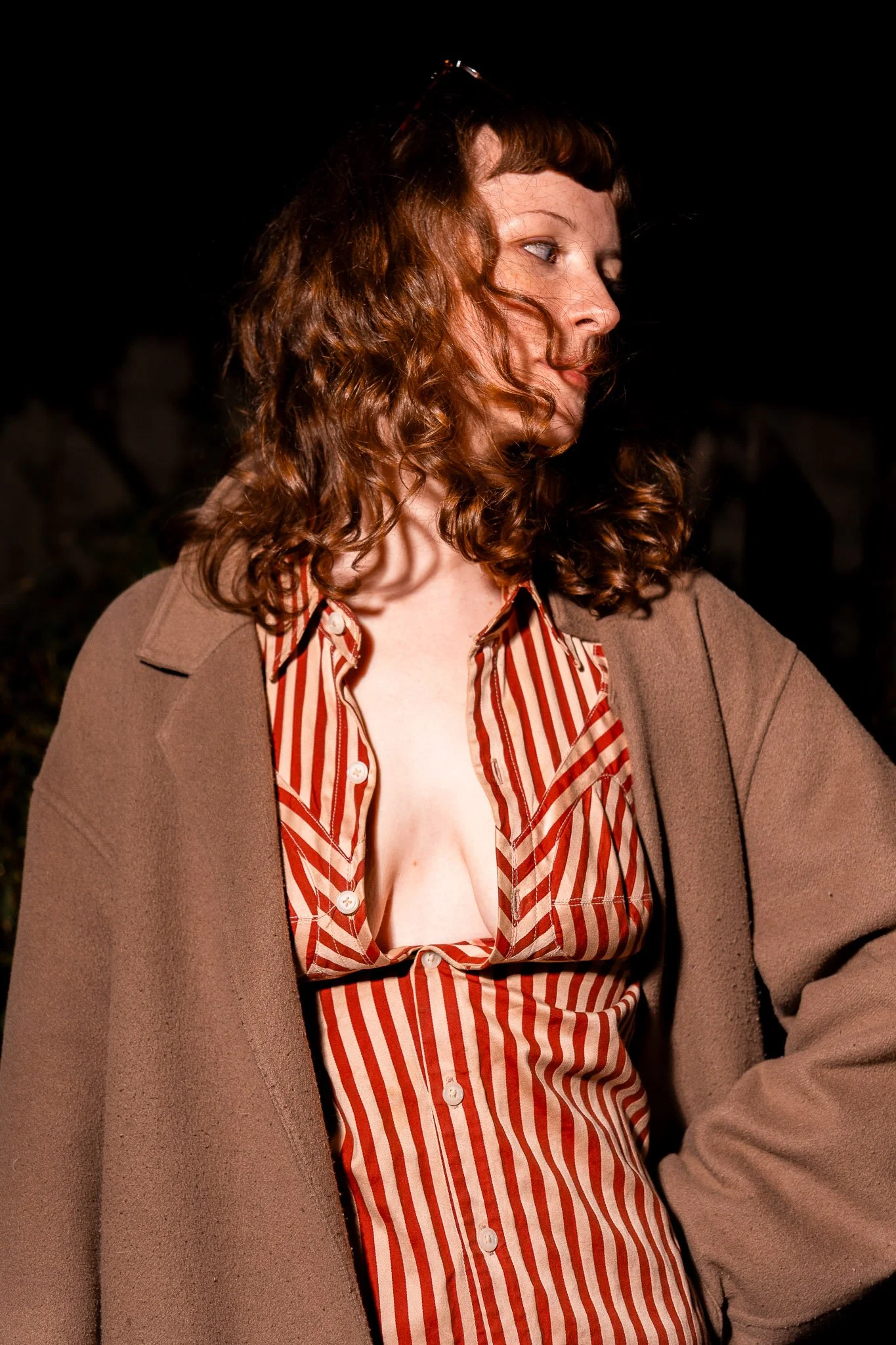 A woman with curly red hair looking to the right, wearing a beige blazer and a red and white striped button-up shirt.