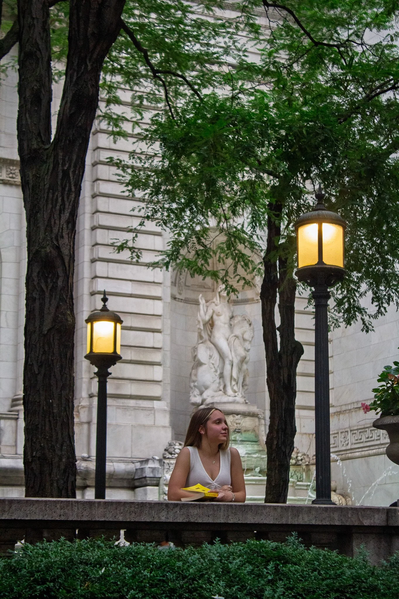 A young woman with long brown hair sits outdoors, holding yellow pamphlets, in front of a fountain and a building with classical sculptures and architectural details. Two street lamps cast warm light, trees with green leaves frame the scene.