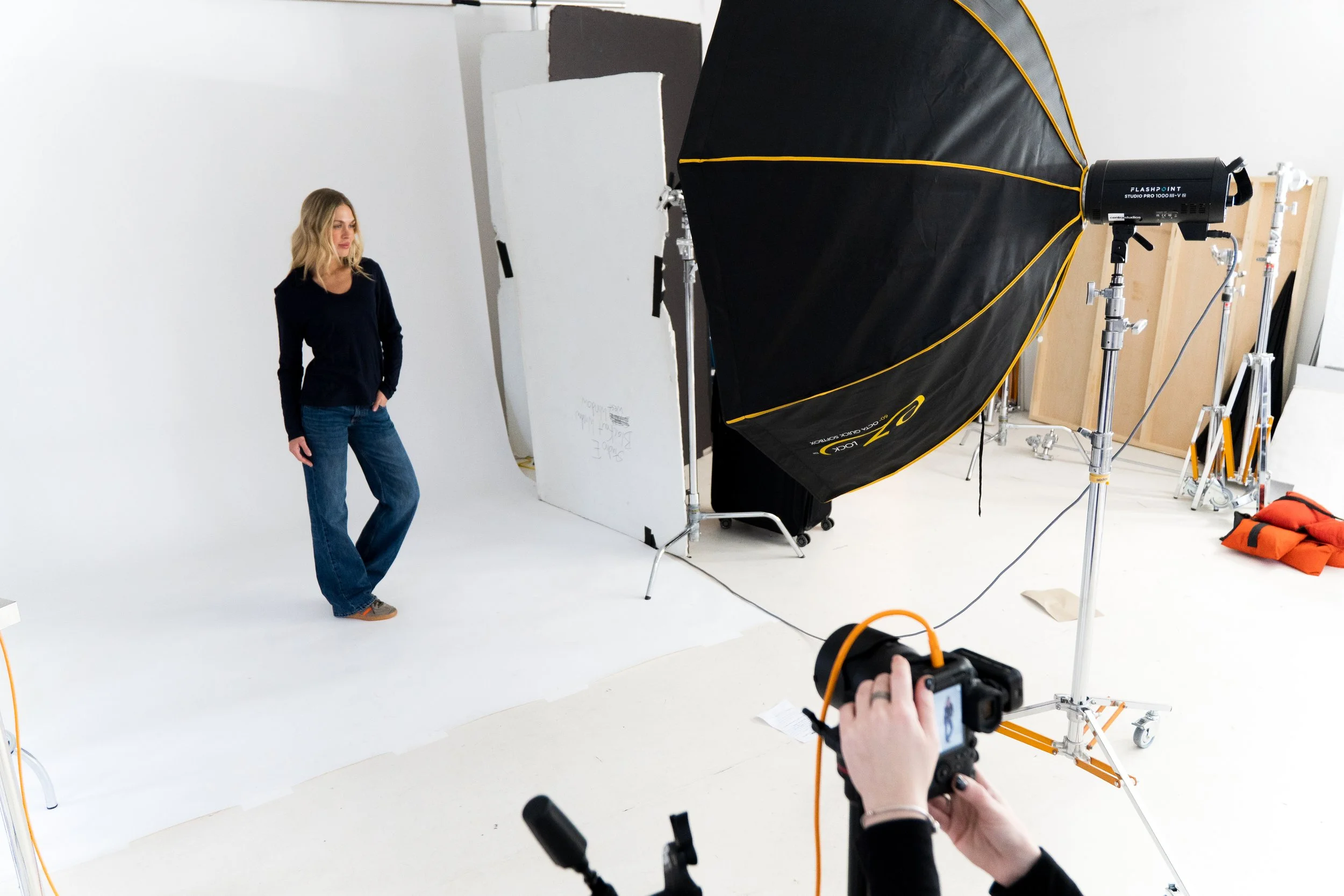 A woman with blonde hair in a black shirt and jeans posing in a photography studio with a large black and yellow umbrella reflector, a white backdrop, and photography equipment around her.
