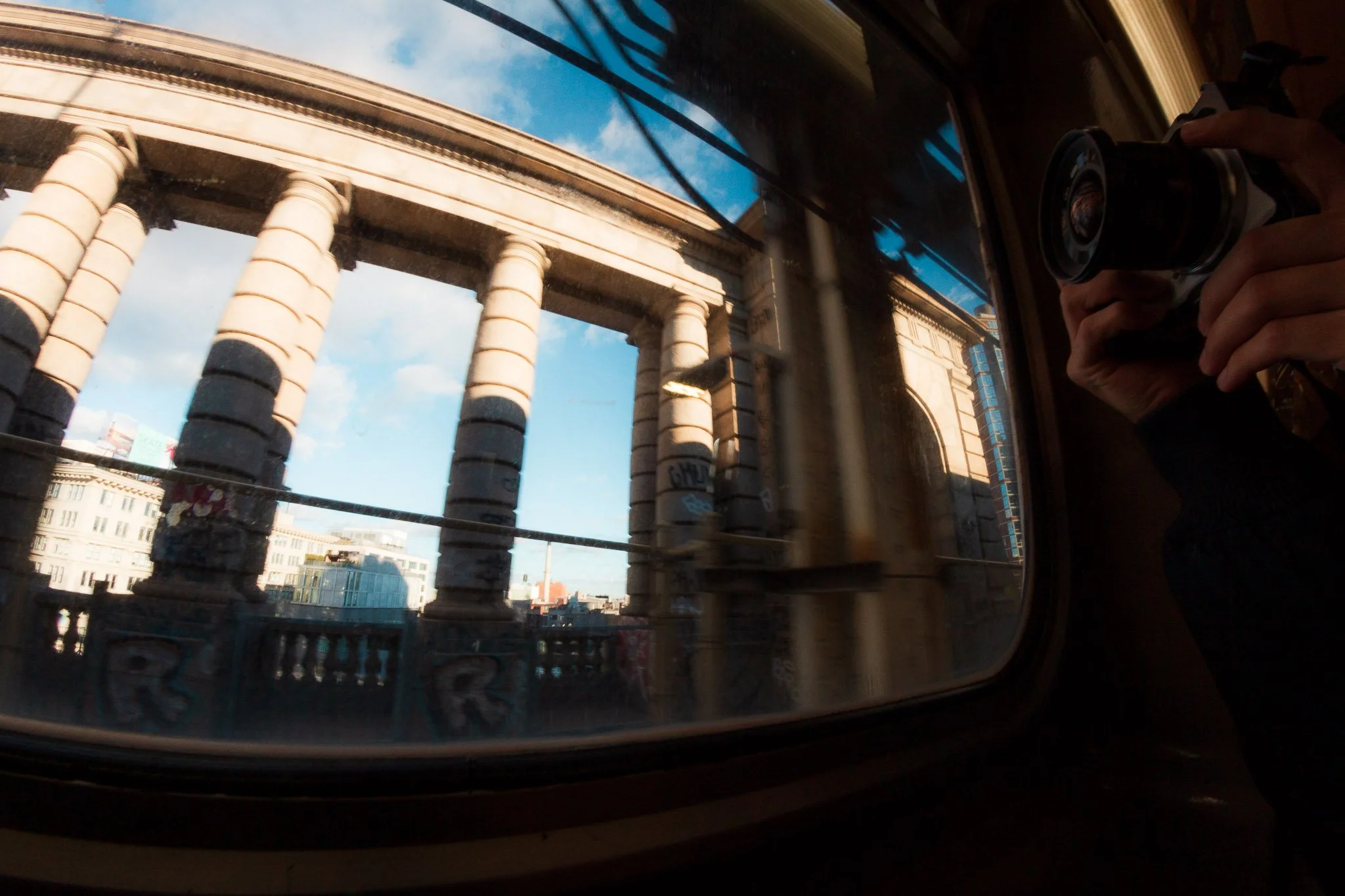 Reflection of a classical-style building with tall columns through a vehicle window, with a person taking a photo using a camera in the upper right corner.