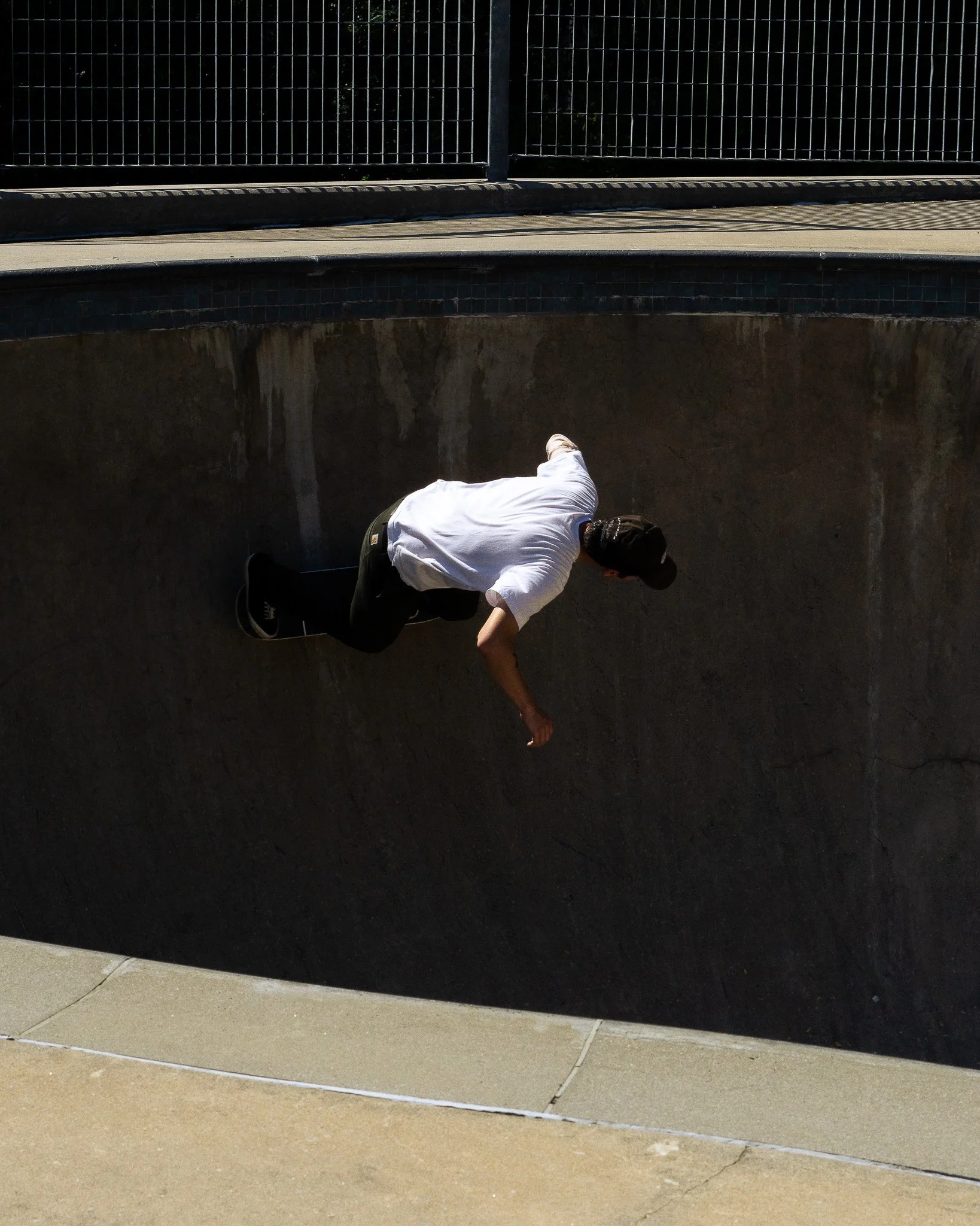 A person wearing a white shirt and black cap is crouched down near a concrete ledge, with their arm extended toward the ground, next to a black metal fence on a sidewalk.