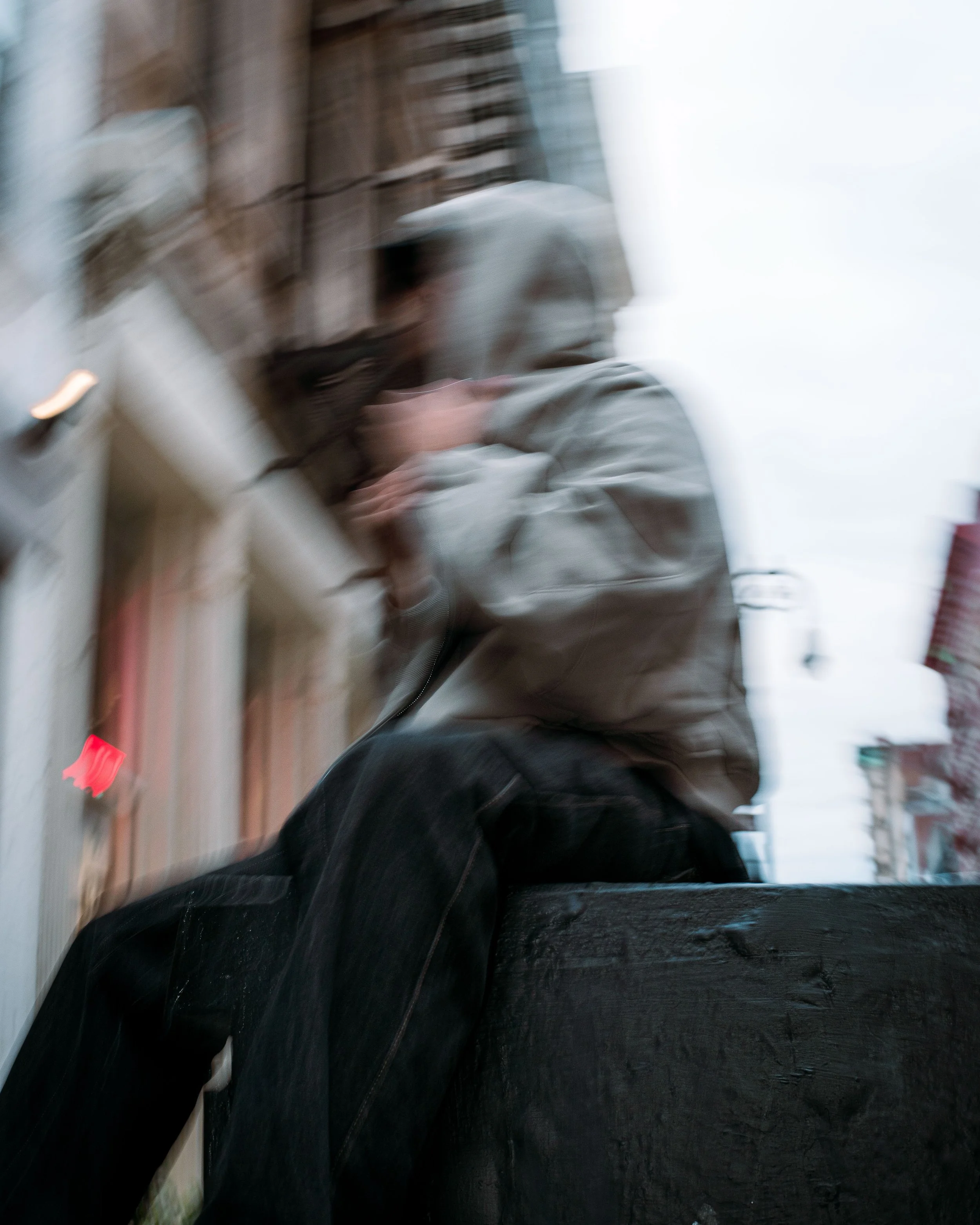 A person wearing a hoodie and dark pants sitting on a black surface, holding a phone near their face, outdoors on a city street with buildings and electrical wiring visible in the background.