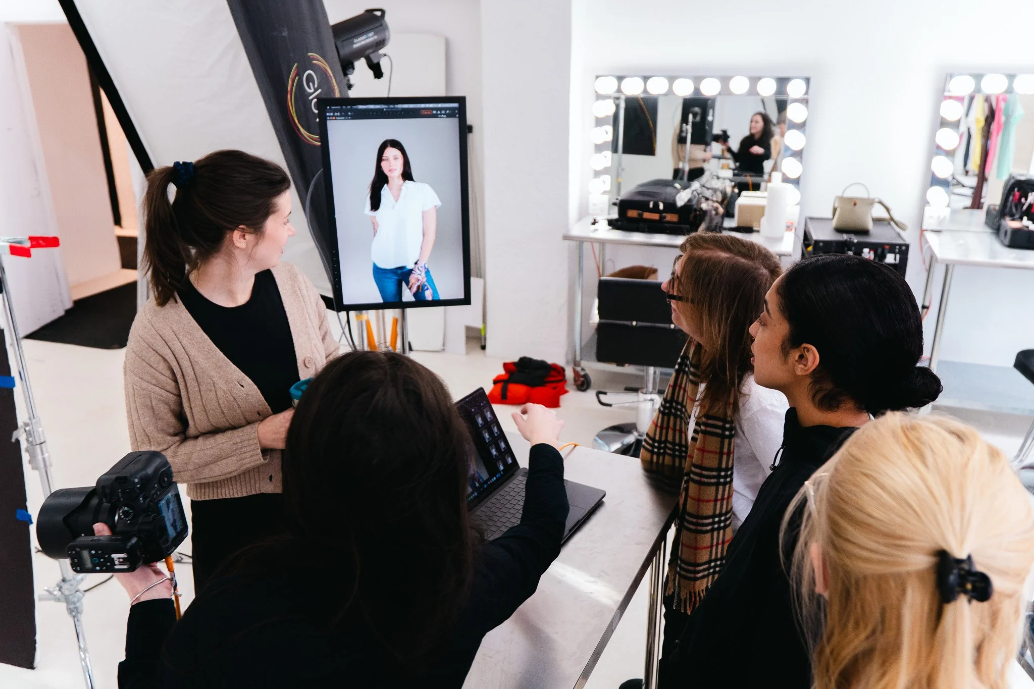 A photography or fashion shoot in a studio with a model on a screen and several women observing, surrounded by cameras, lighting, and makeup mirrors.