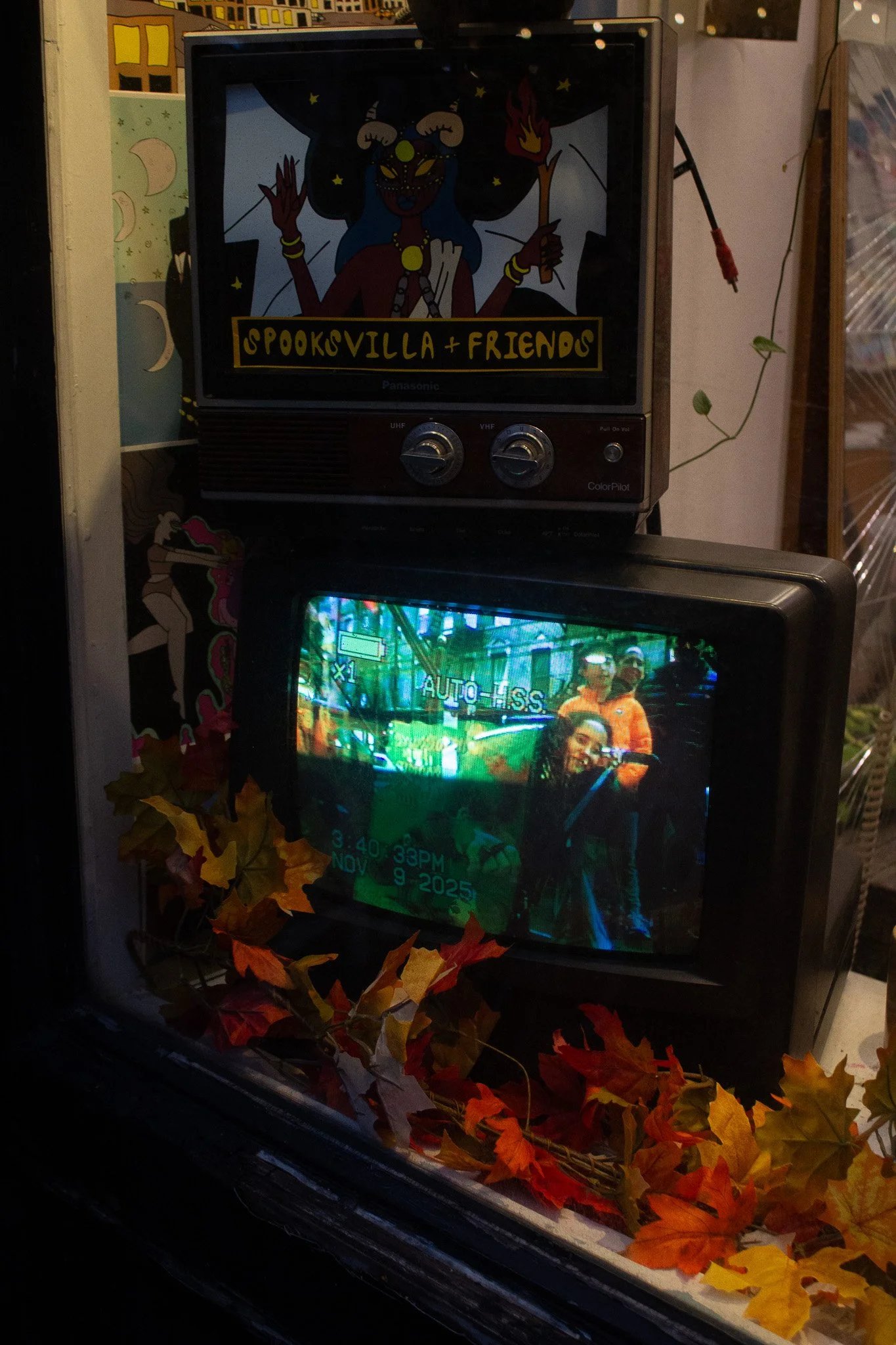 An old TV showing a group of people outdoors with trees in the background, and a vintage camera overlay. On top of it is another older TV with artwork titled 'Spooksville + Friends,' featuring a cartoon character holding a torch. Autumn leaves decora