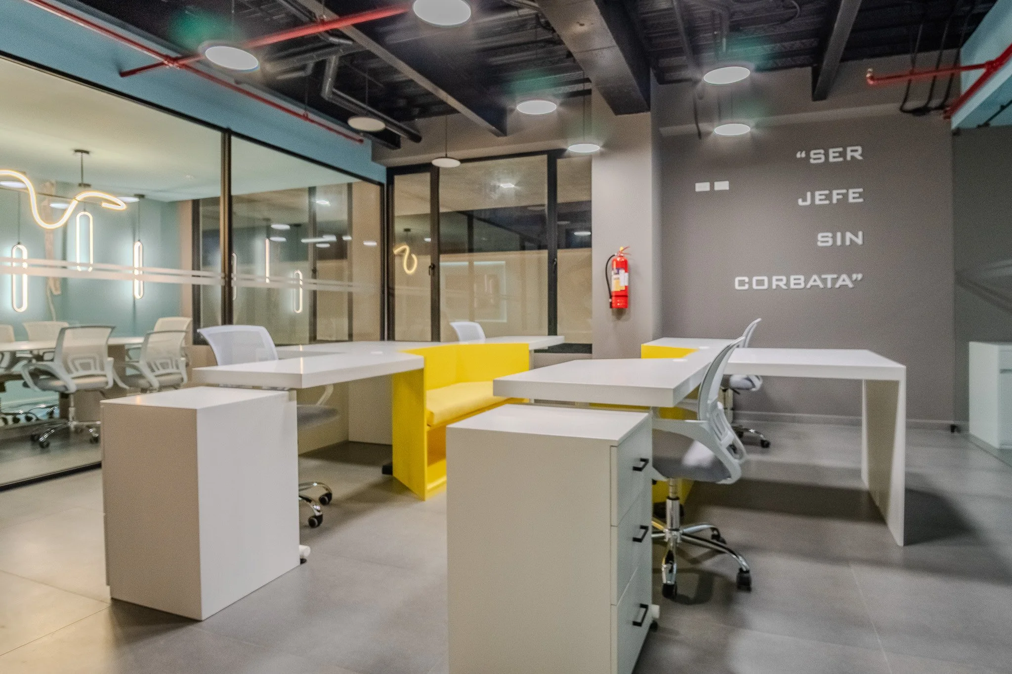 Modern office with multiple white desks, white office chairs, and yellow accents, separated by glass walls with a quote on the gray wall reading 'SER JEFES SIN GORBATA'.