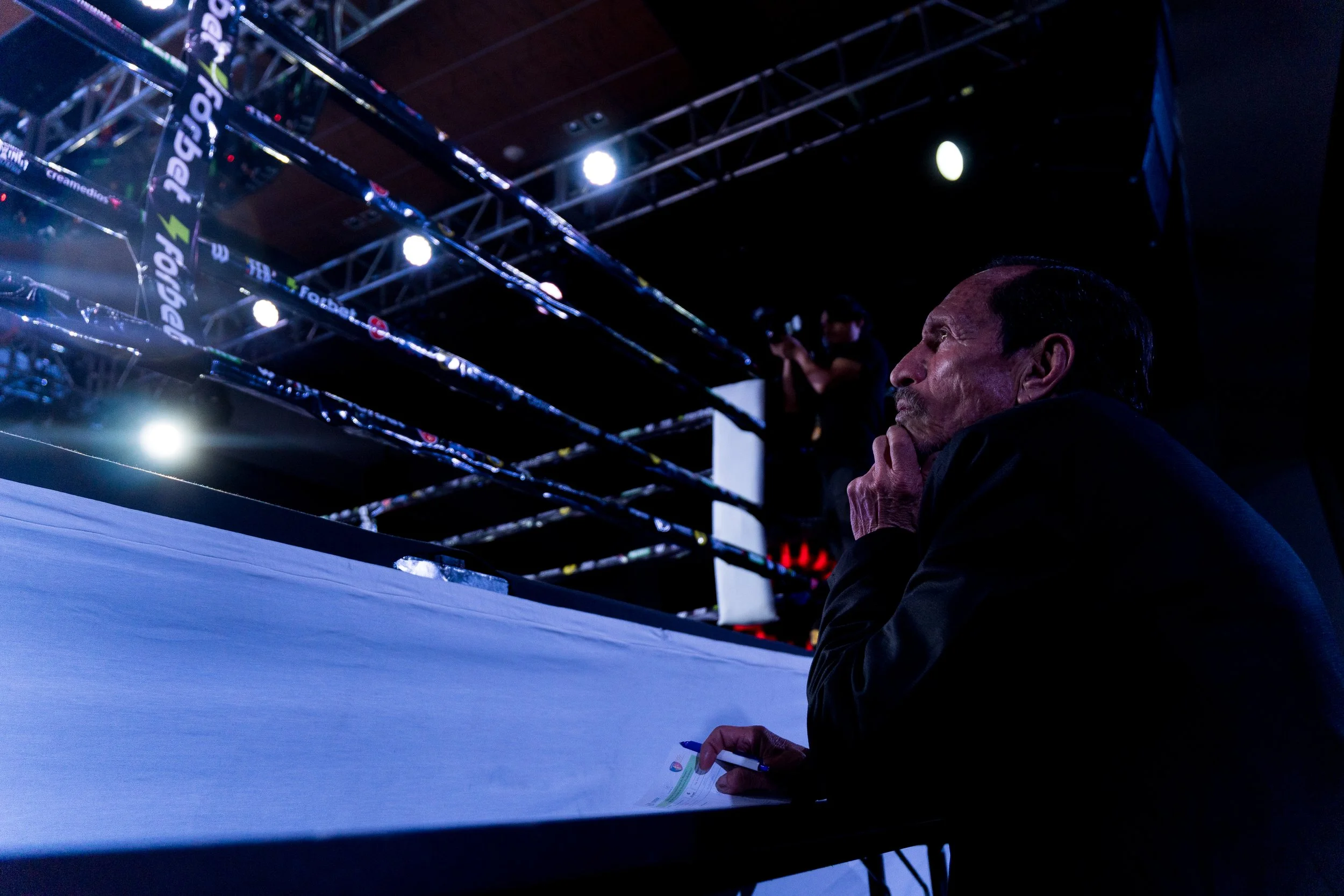A man sitting in an arena or stadium, looking thoughtful or pensive, with numbered boxing or wrestling rings in the background and a photographer capturing the event.
