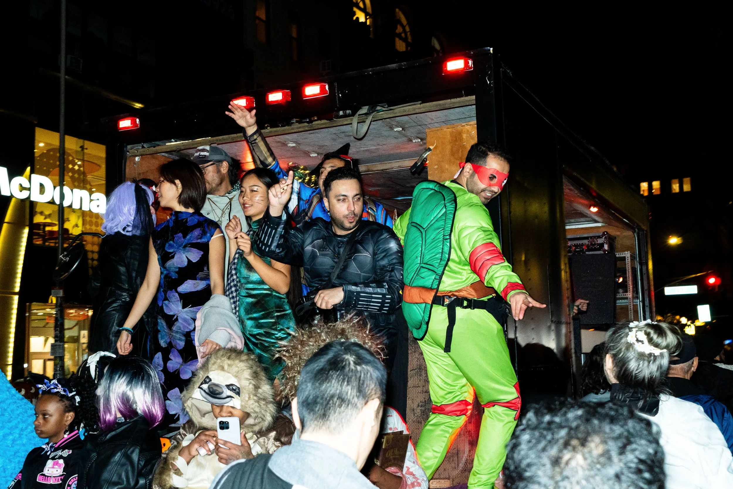People dressed in costumes on a parade float at night, with some taking photos and others dancing or posing.