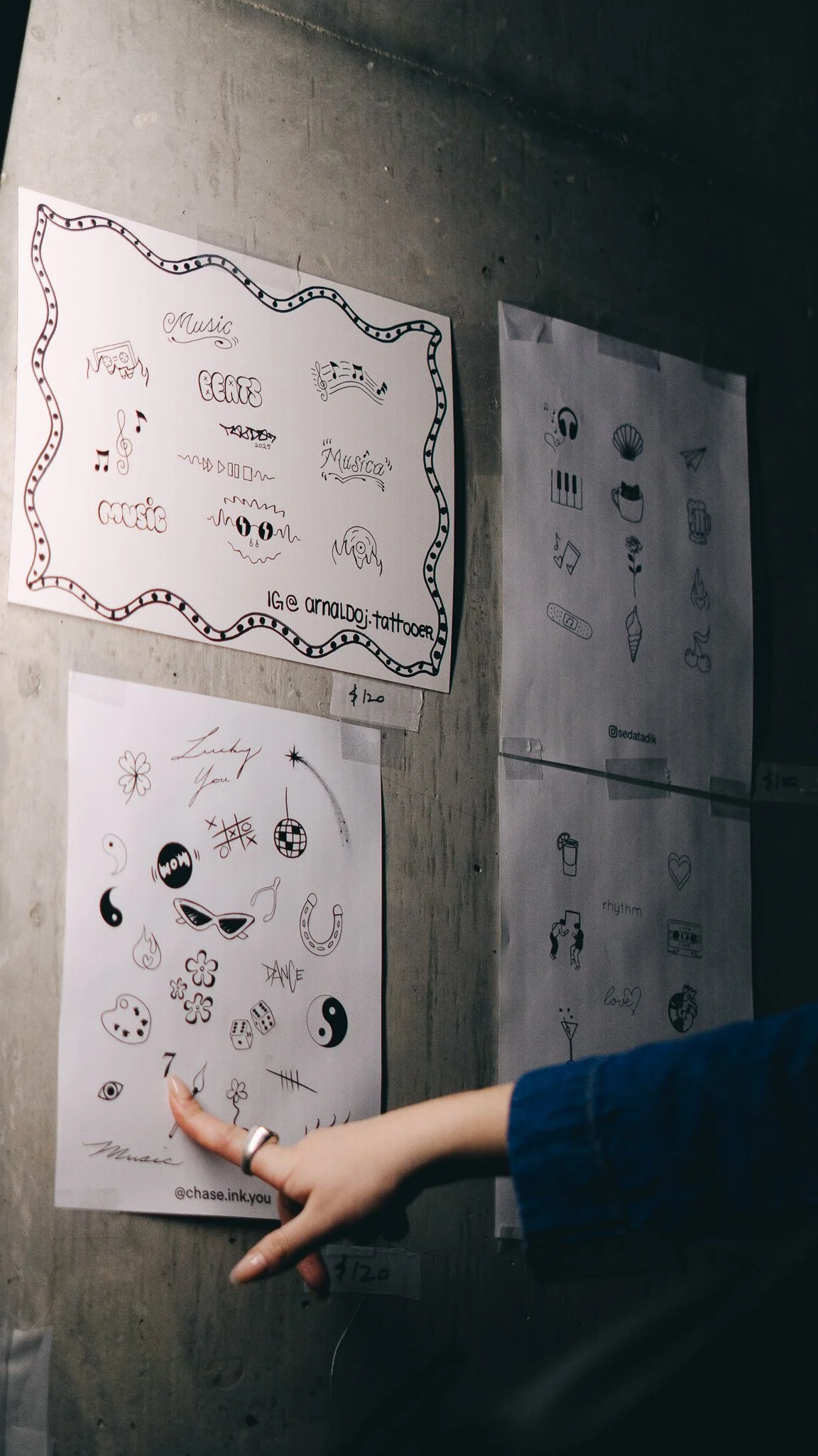 A person's hand pointing to a sheet of temporary tattoos with various black and white designs, including smiley faces, music notes, a disco ball, flowers, and other symbols, held against a concrete wall.