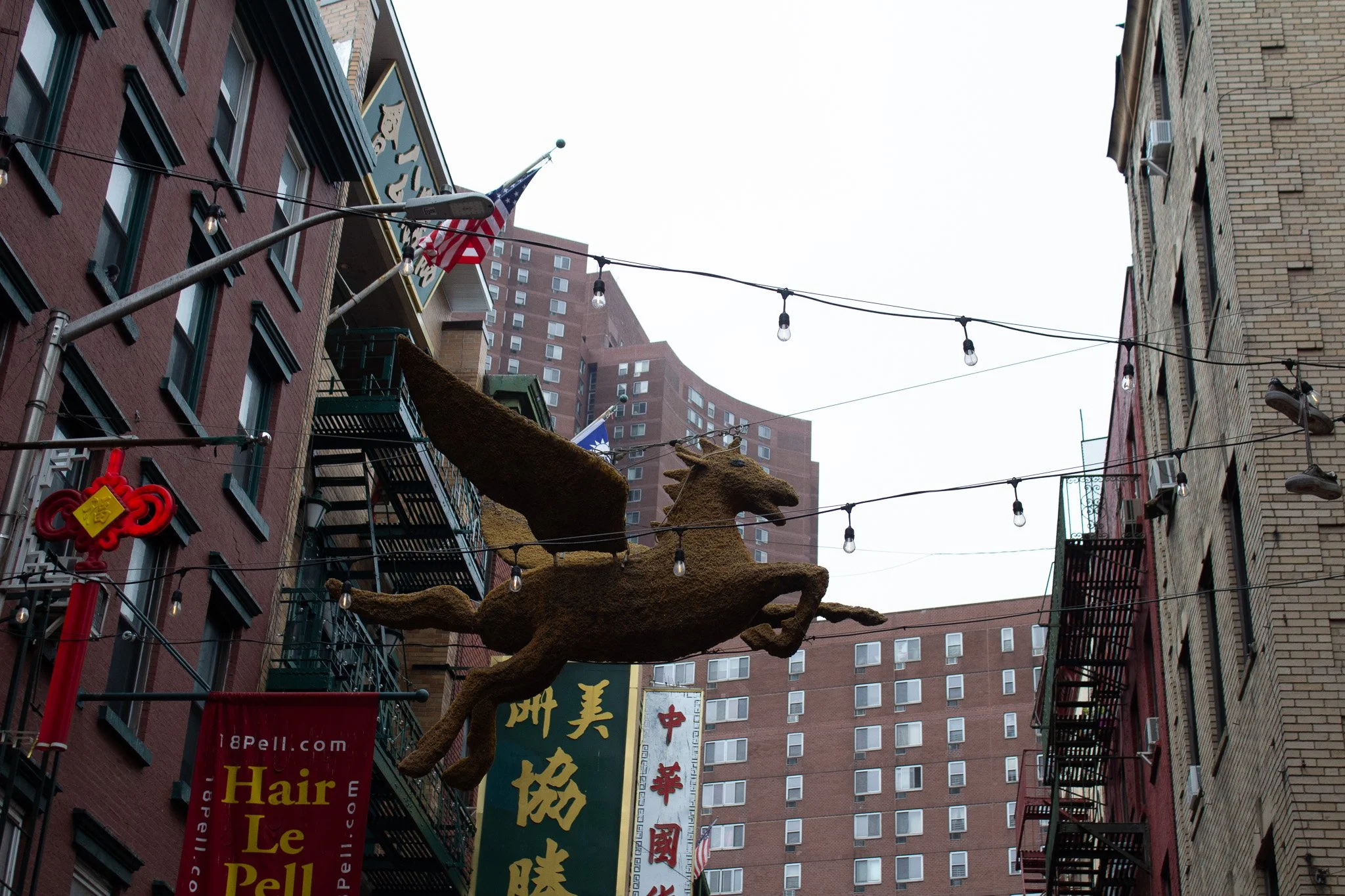 Decorative flying horse sculpture hanging above a city street with buildings, string lights, and American flag.