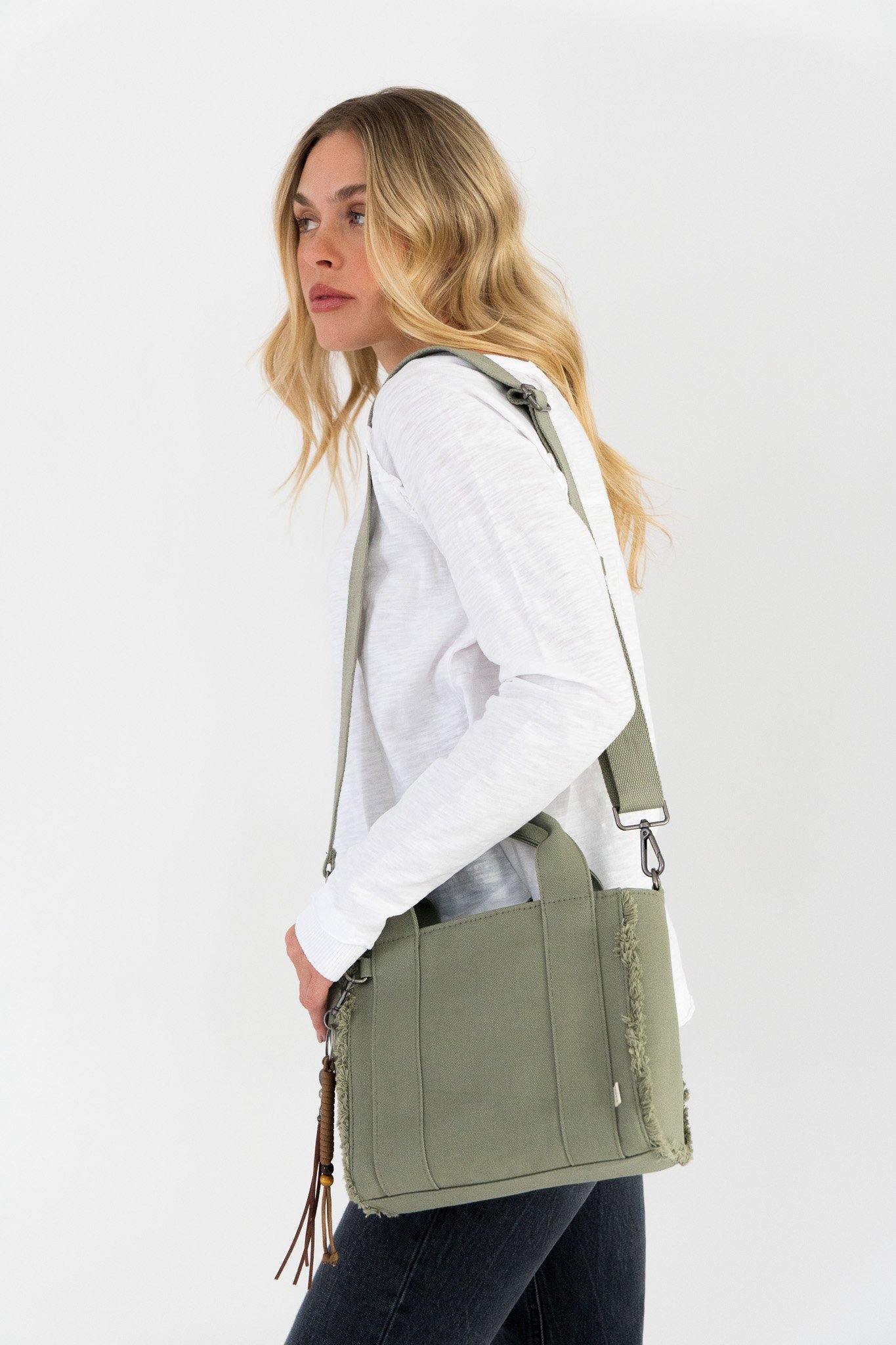 A woman with long, wavy blonde hair wearing a white long-sleeve shirt and black pants, carrying a large green tote bag over her shoulder, standing against a plain white wall.