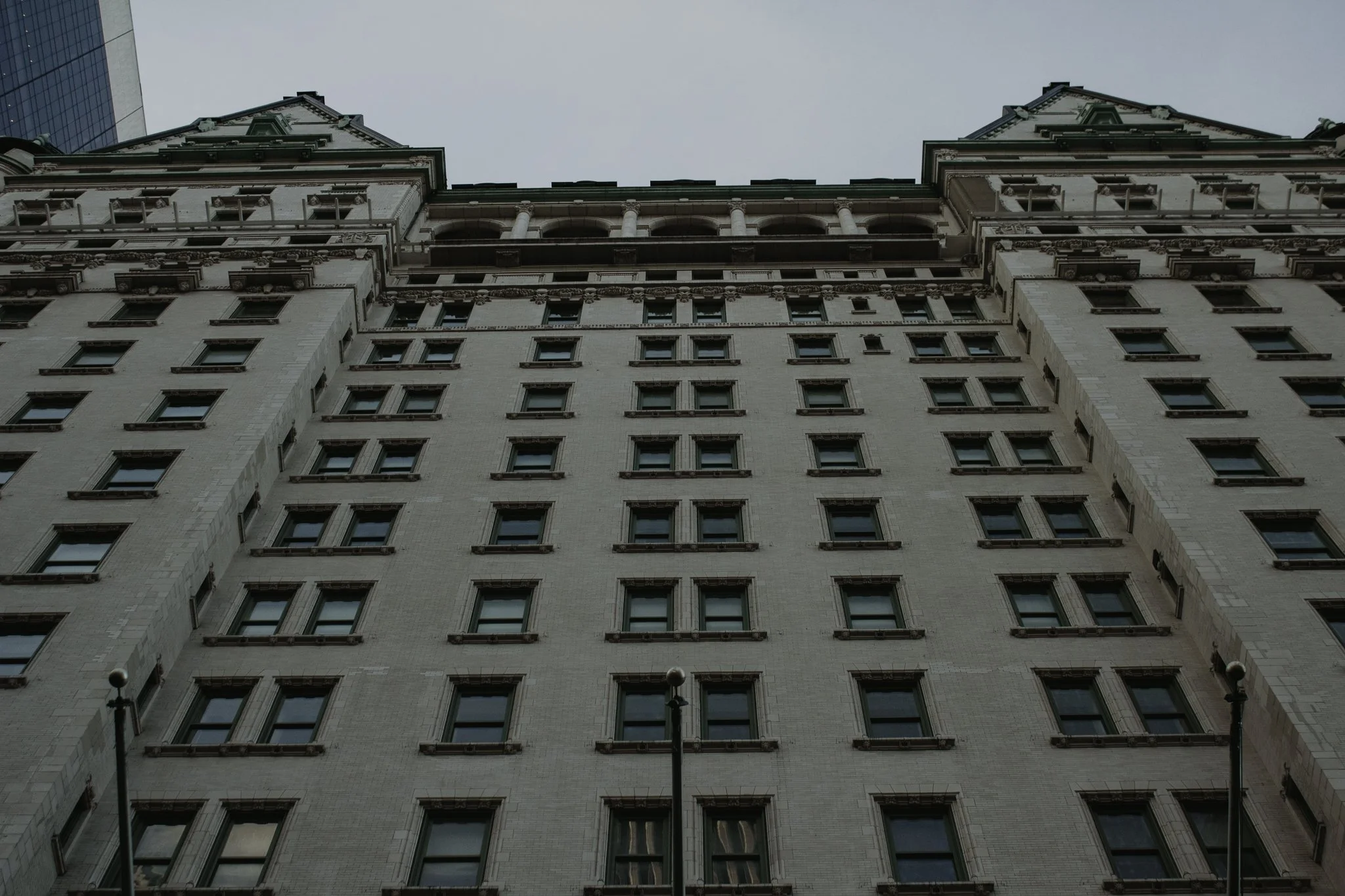 Looking up at an ornate multi-story building with a classical architectural style, including detailed window frames, columns, and decorative trim, with a modern glass building visible on the left side and street lamps in the foreground.