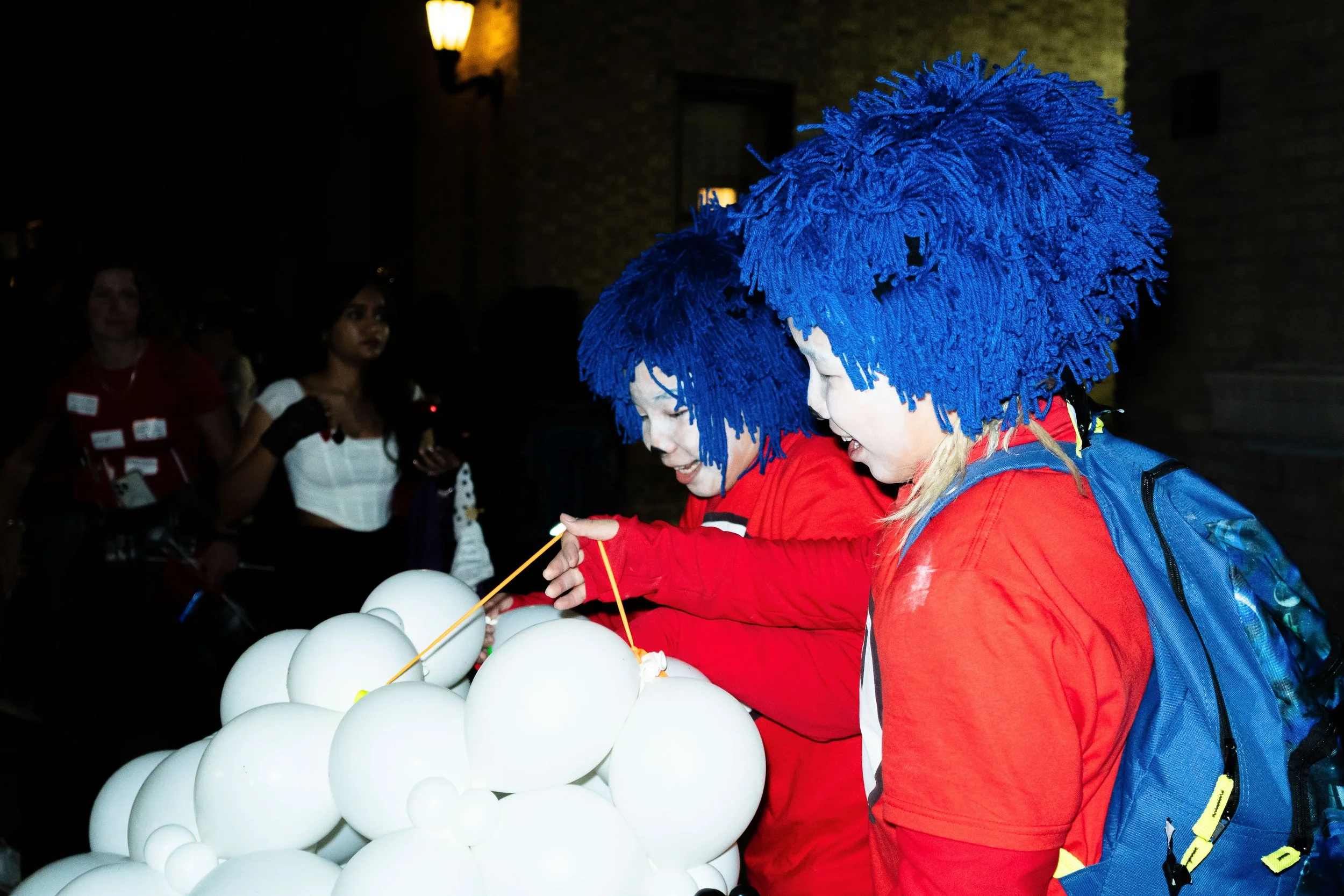 Two children in blue wigs, red shirts, and backpacks are making a sculpture with white balloons in a dimly lit room, while other people watch in the background.