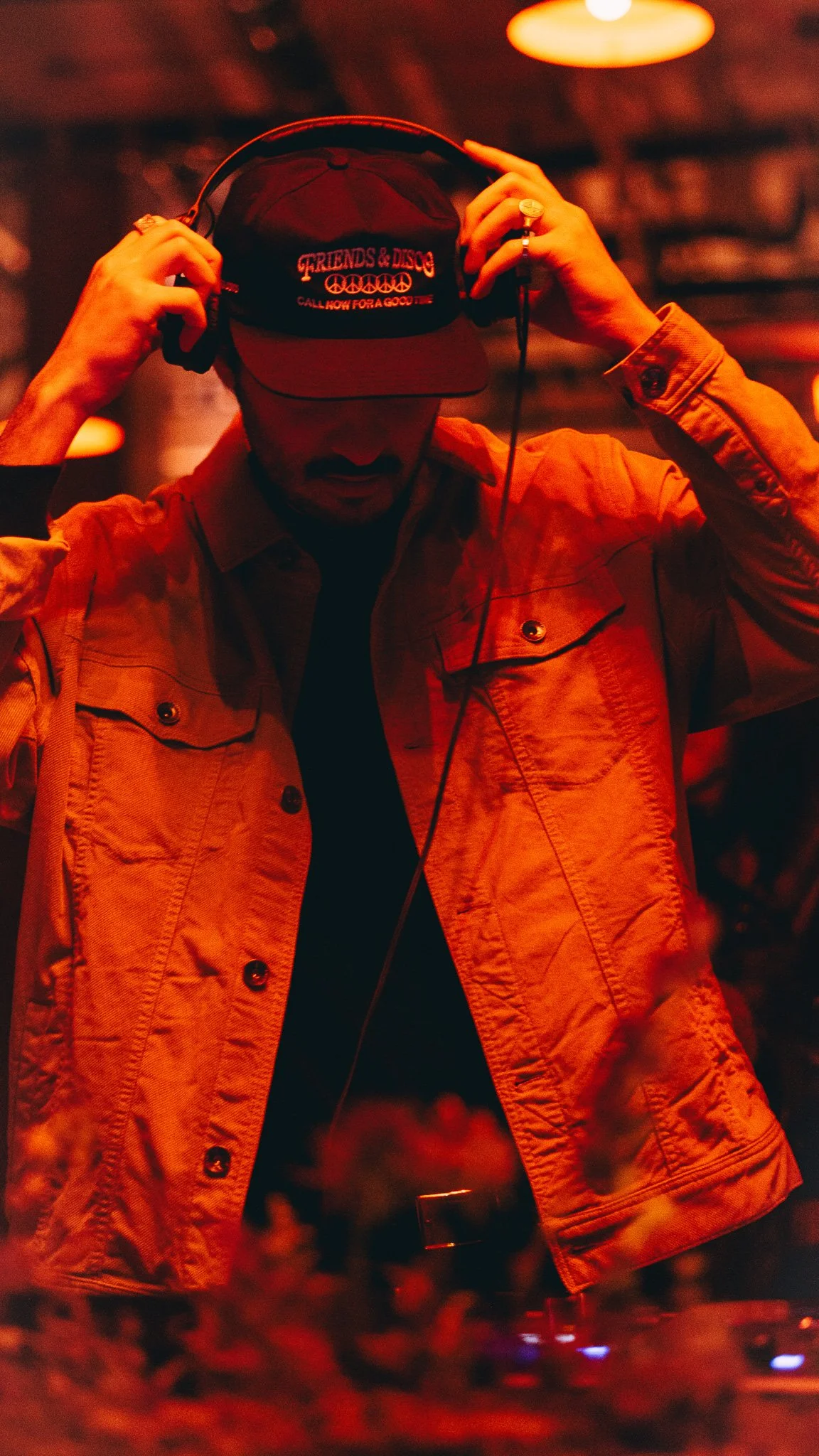 Person wearing a cap and headphones adjusting them, with a dimly lit, red-tinged background, possibly at a nightclub or music event.