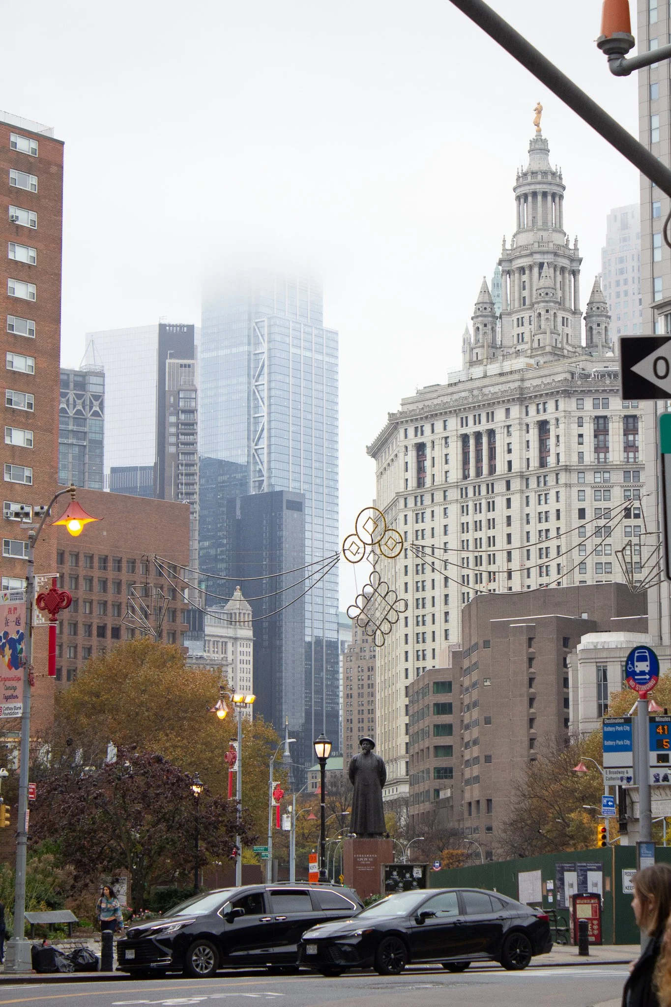 City street scene with tall skyscrapers, historic buildings, street decorations, trees, streetlights, cars, and pedestrians.