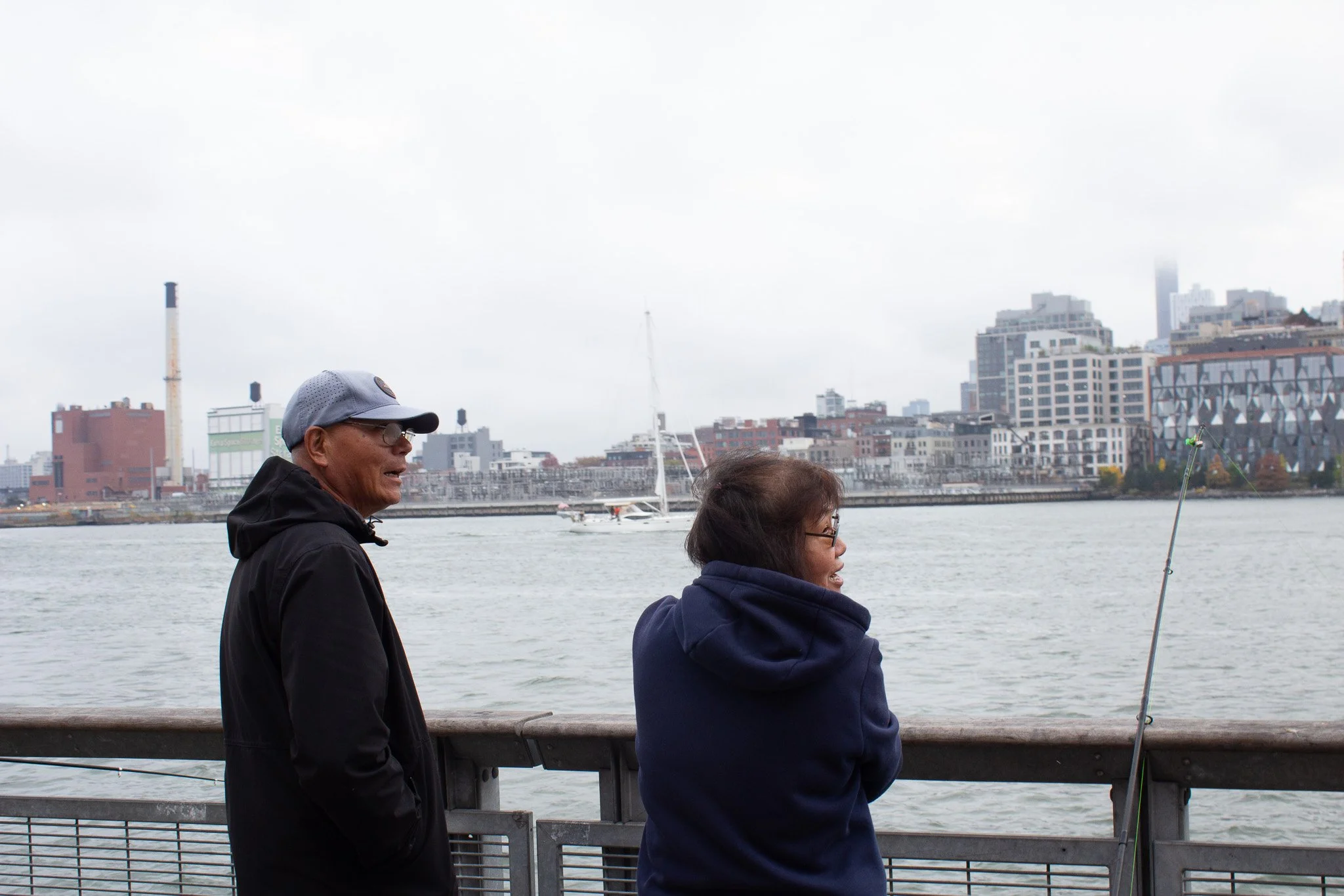 A man and woman standing on a waterfront promenade, with buildings and a sailboat in the background, as the woman fishes with a rod.