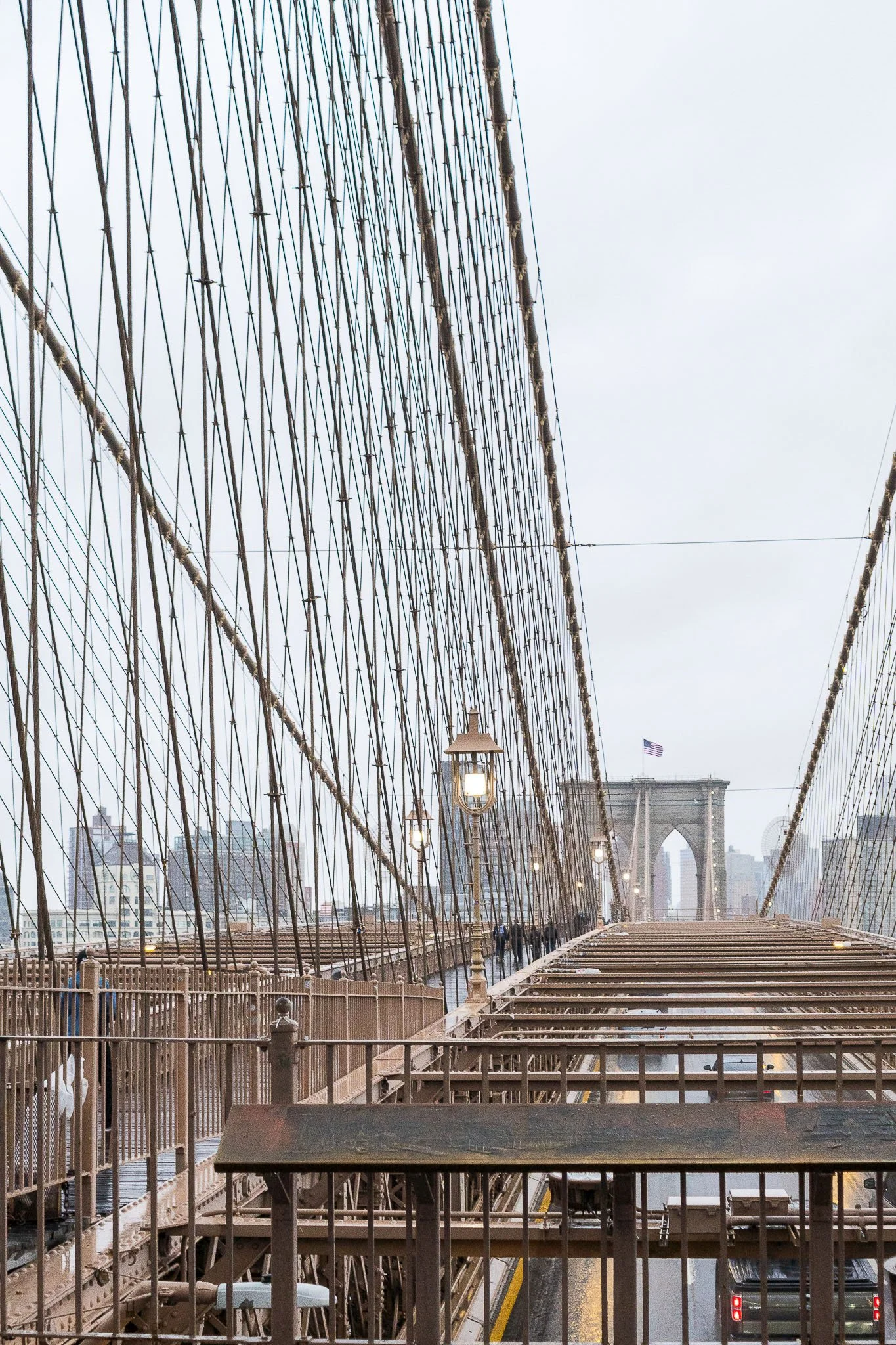 View of Brooklyn Bridge in New York City with a cloudy sky, featuring the cables and the main tower with an American flag, and a walkway with lamps and people walking.