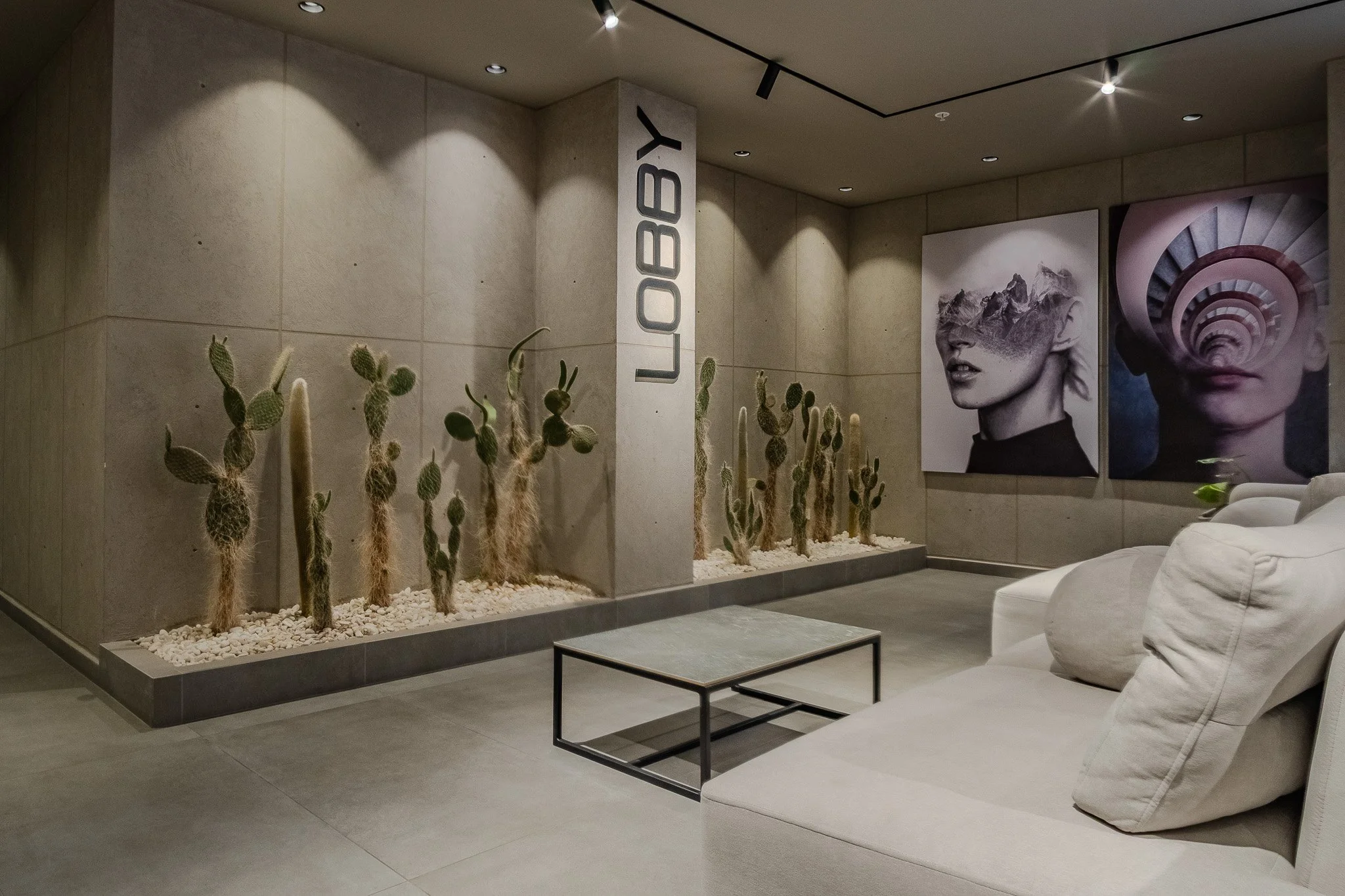 Modern interior space with a beige wall, desert cacti in a bed of white stones, and large black-and-white art posters, including one of a woman with mountains in her hair and another with a spiral staircase pattern, with a white sofa and black metal 