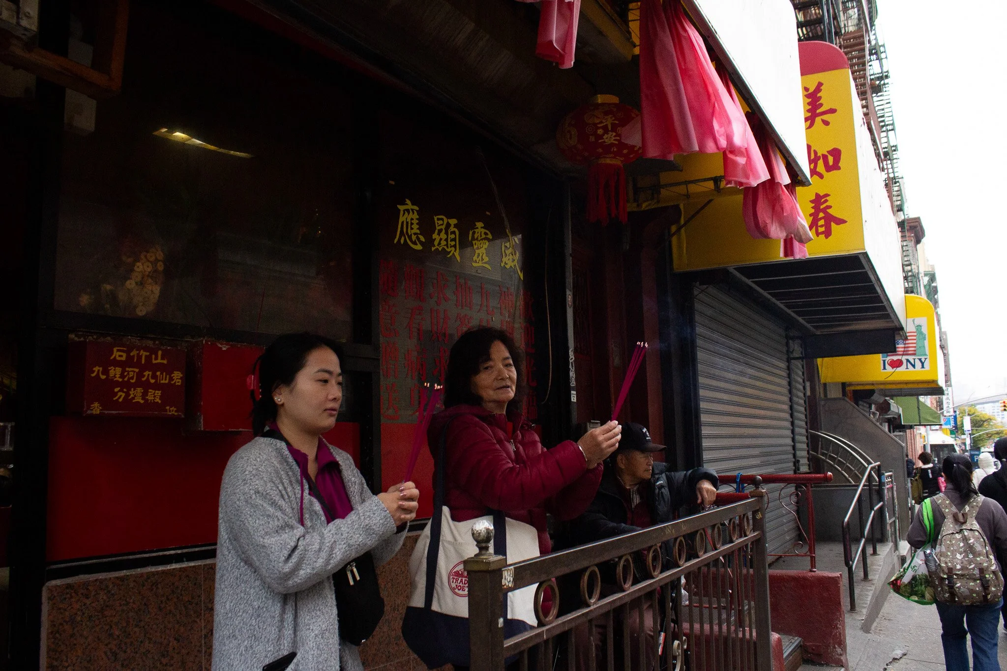 Three women standing on a sidewalk in front of a store, holding pink incense sticks. They are dressed in casual clothing and appear to be in prayer or paying respects. The store has red and yellow signage with Chinese characters, and pink decorations