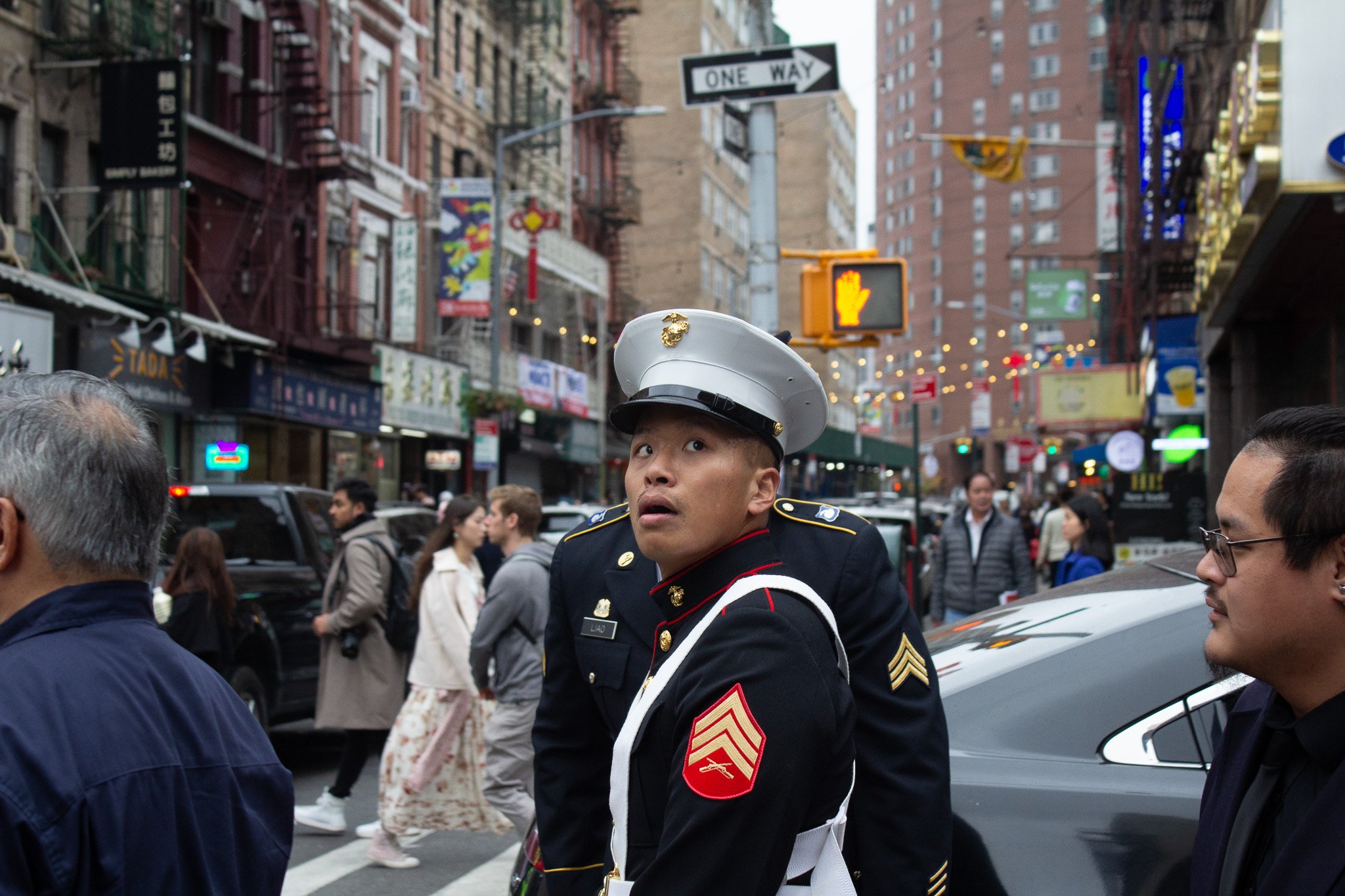A street scene in an urban area with a uniformed member of the military or police in the foreground, looking surprised or confused. There are several pedestrians walking in the background, tall buildings, street signs, and colorful lights hanging acr