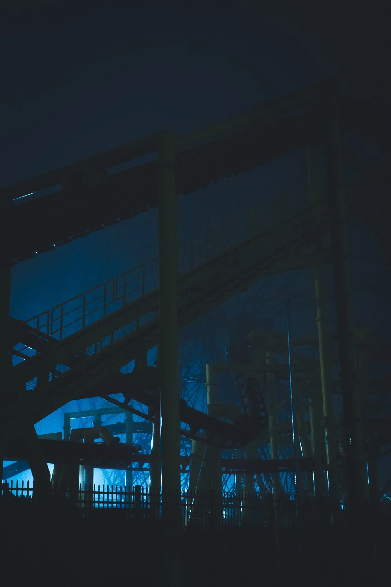 Dark amusement park or fairground ride with metal structures, appears to be nighttime, faint blue lighting illuminating parts of the rides.