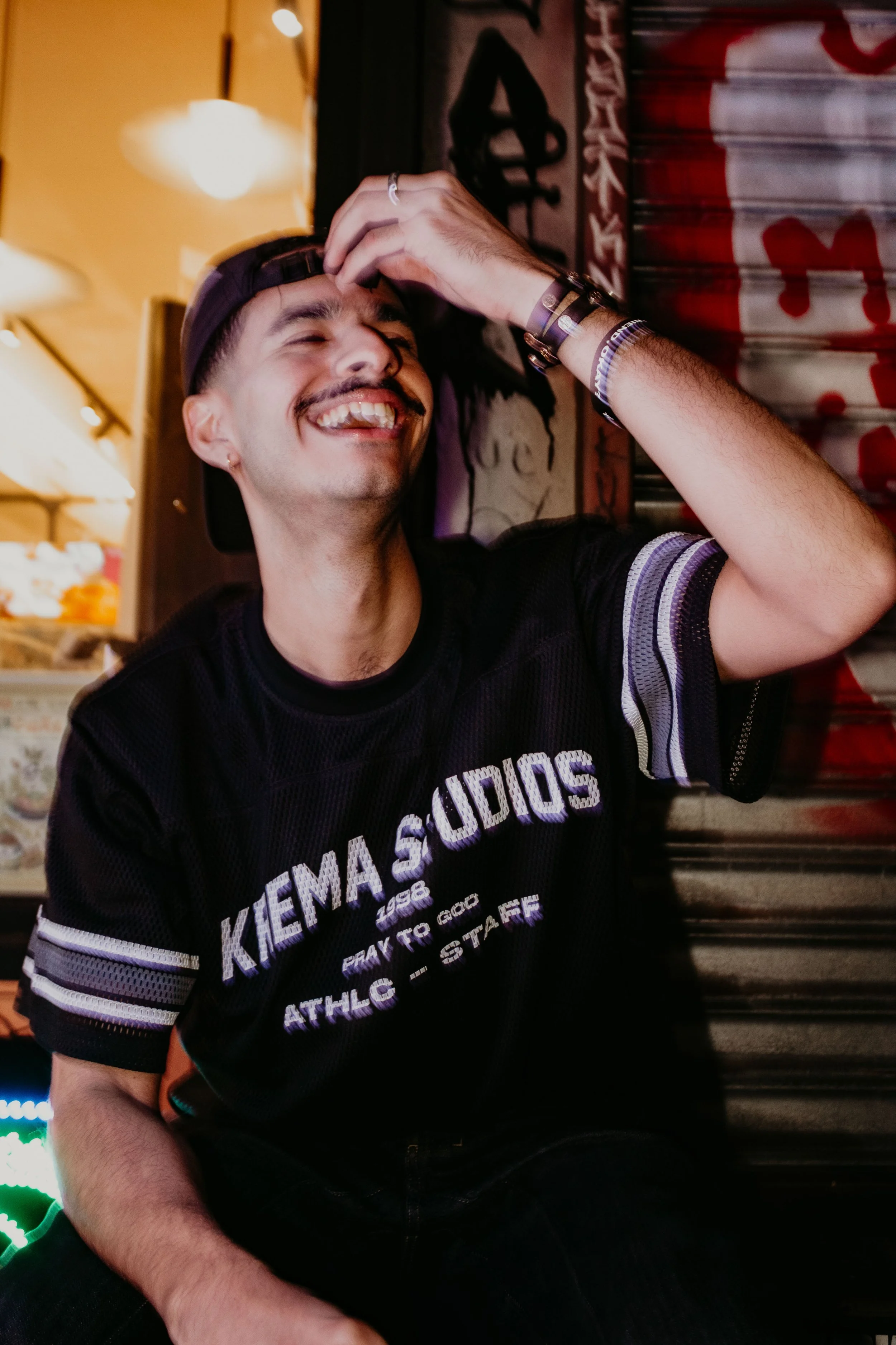 A young man with a mustache and beard, wearing a black baseball cap backwards and a black sports jersey with white and purple lettering, is smiling and laughing while touching his forehead. He is sitting in a dimly lit, colorful room with graffiti an