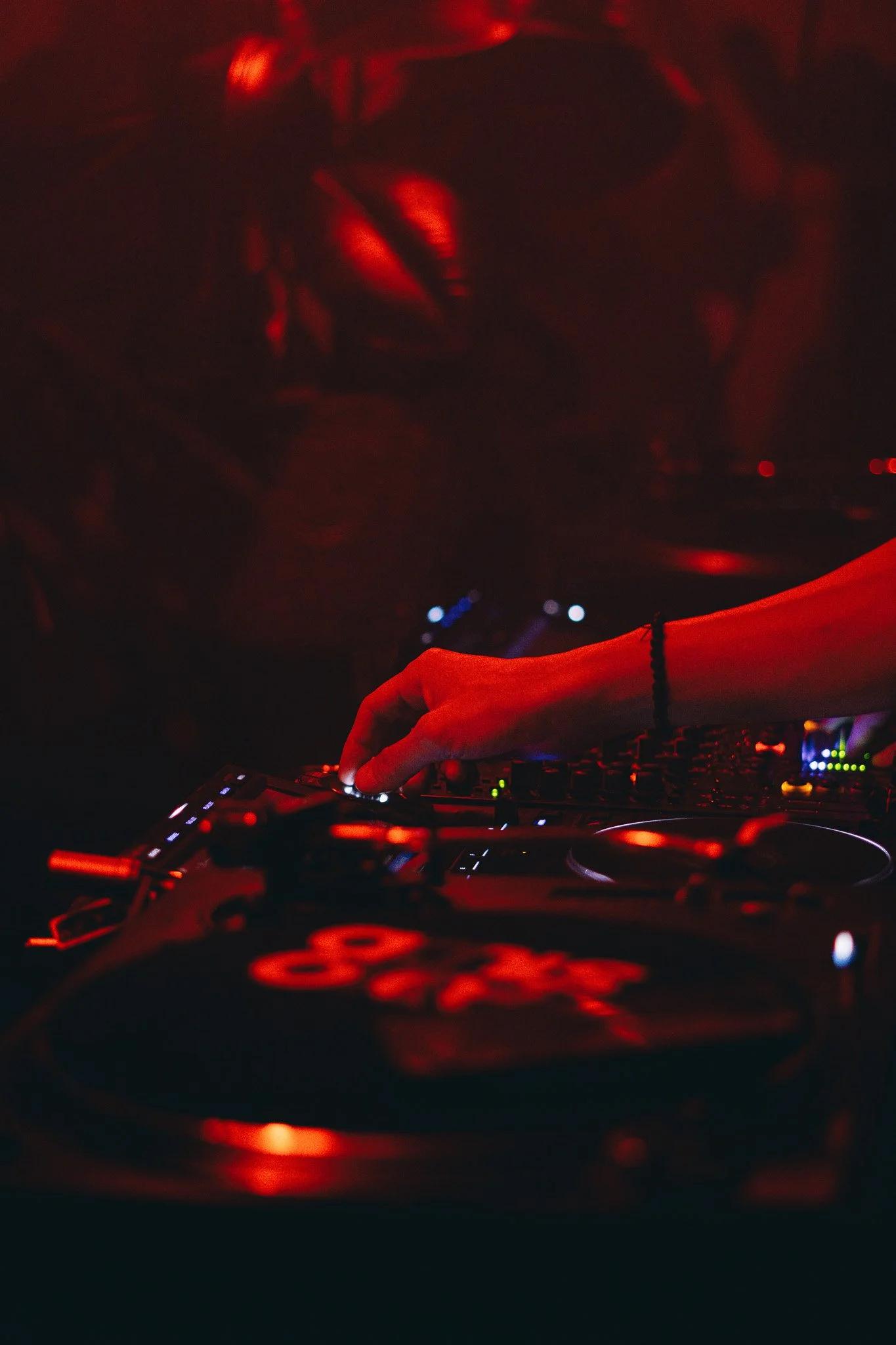 A DJ's hand operating turntables in a dark club with red lighting.