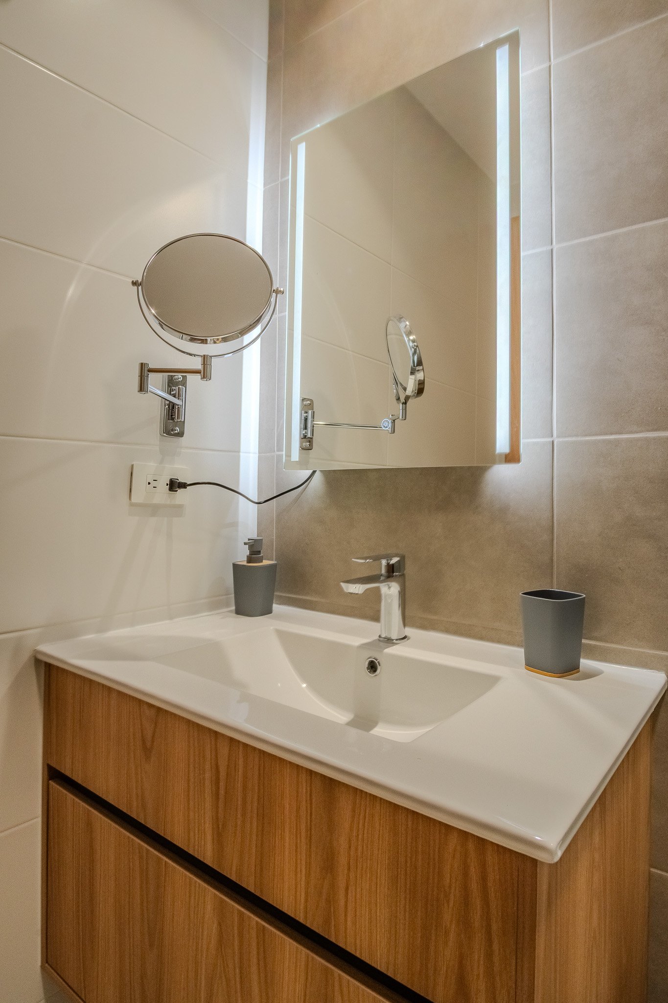 Bathroom vanity with a sink, chrome faucet, dark gray cup, soap dispenser, large mirror, makeup mirror, and beige tiled wall.
