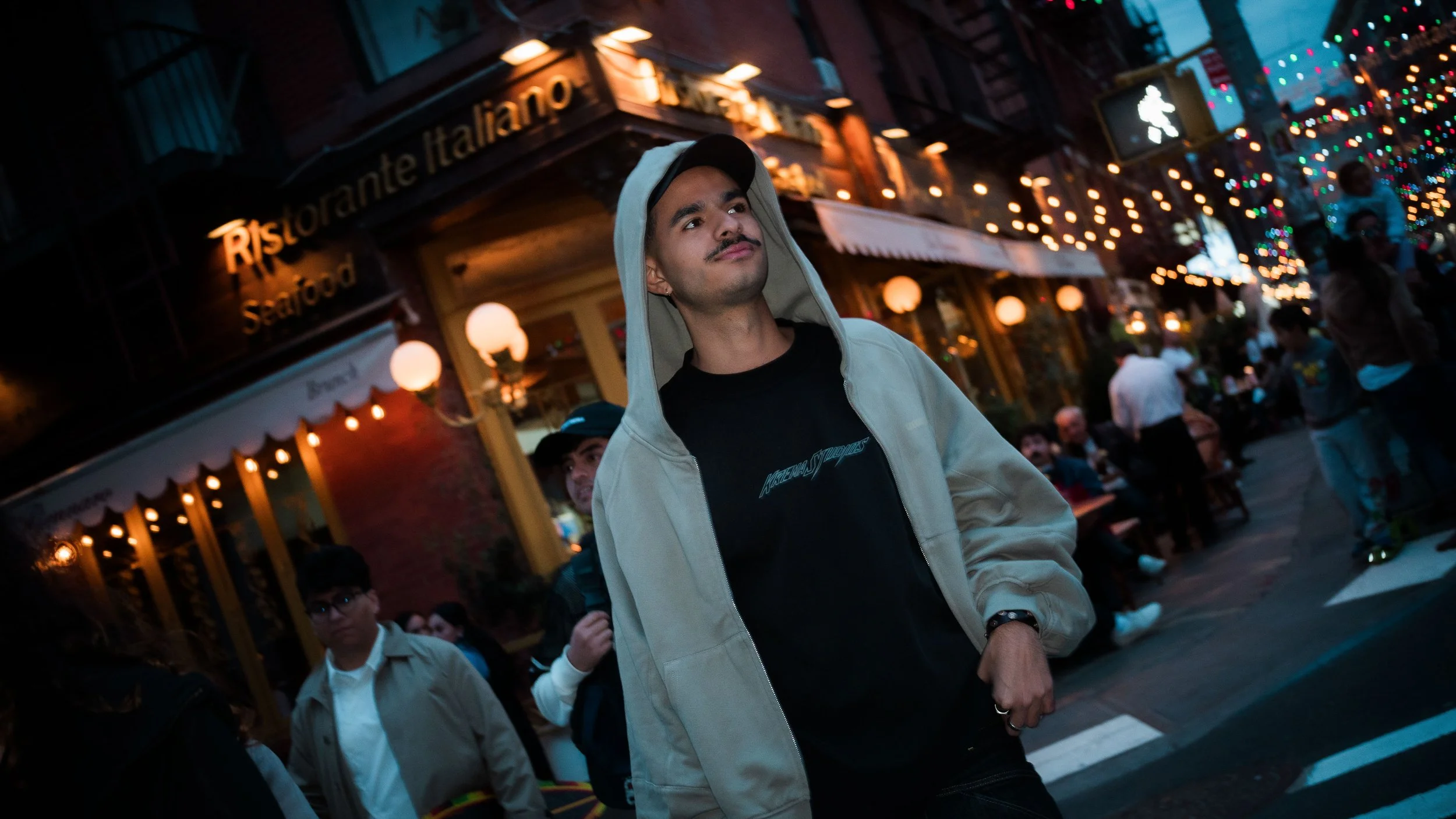 A young man with a mustache in a black t-shirt and beige hoodie walking through a lively street at night, illuminated by decorative lights. Restaurant signs are visible in the background, and other people are seen walking and sitting outside.