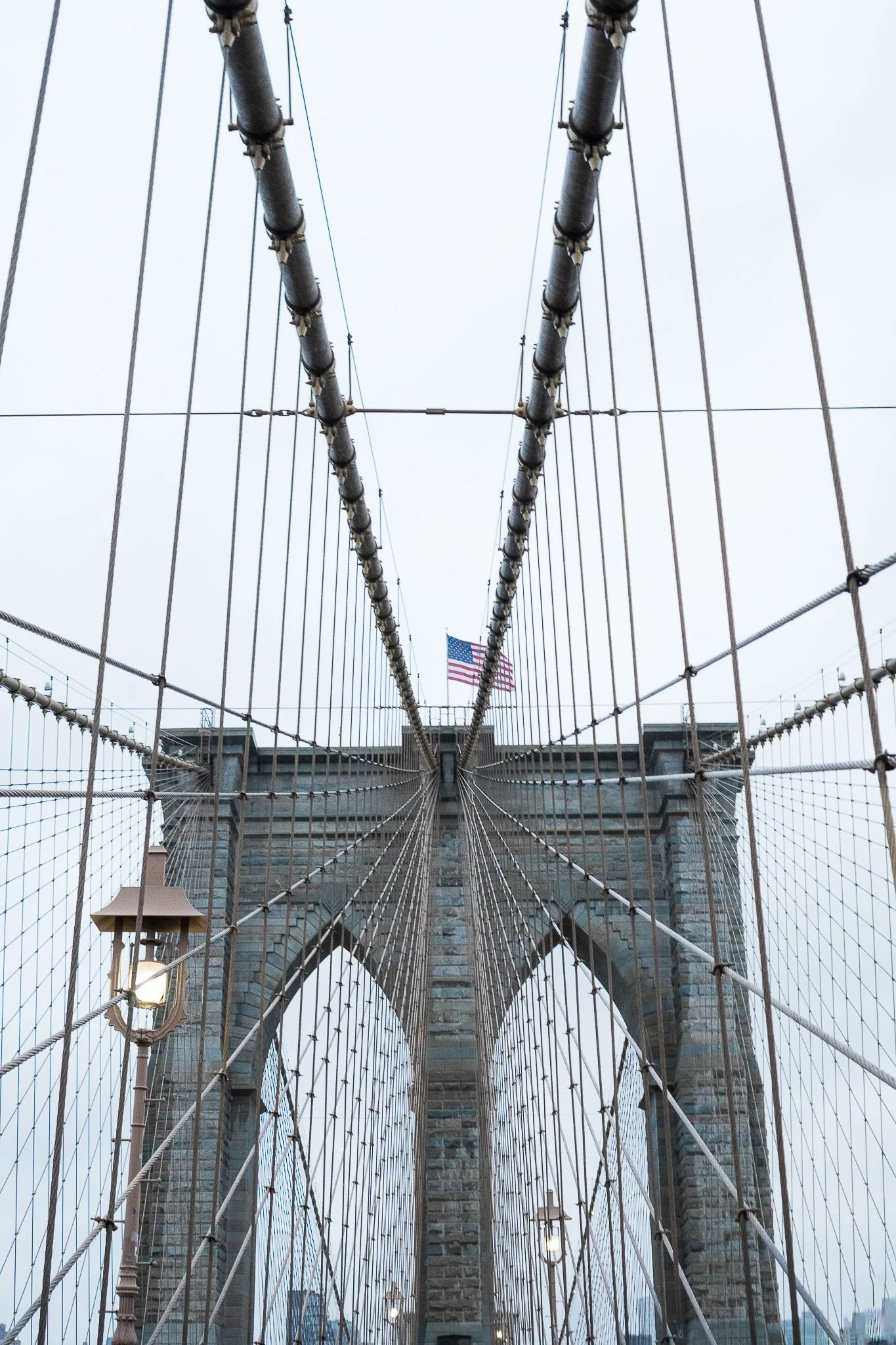 View from the Brooklyn Bridge looking up at the stone towers and suspension cables, with the American flag in the background on a cloudy day.